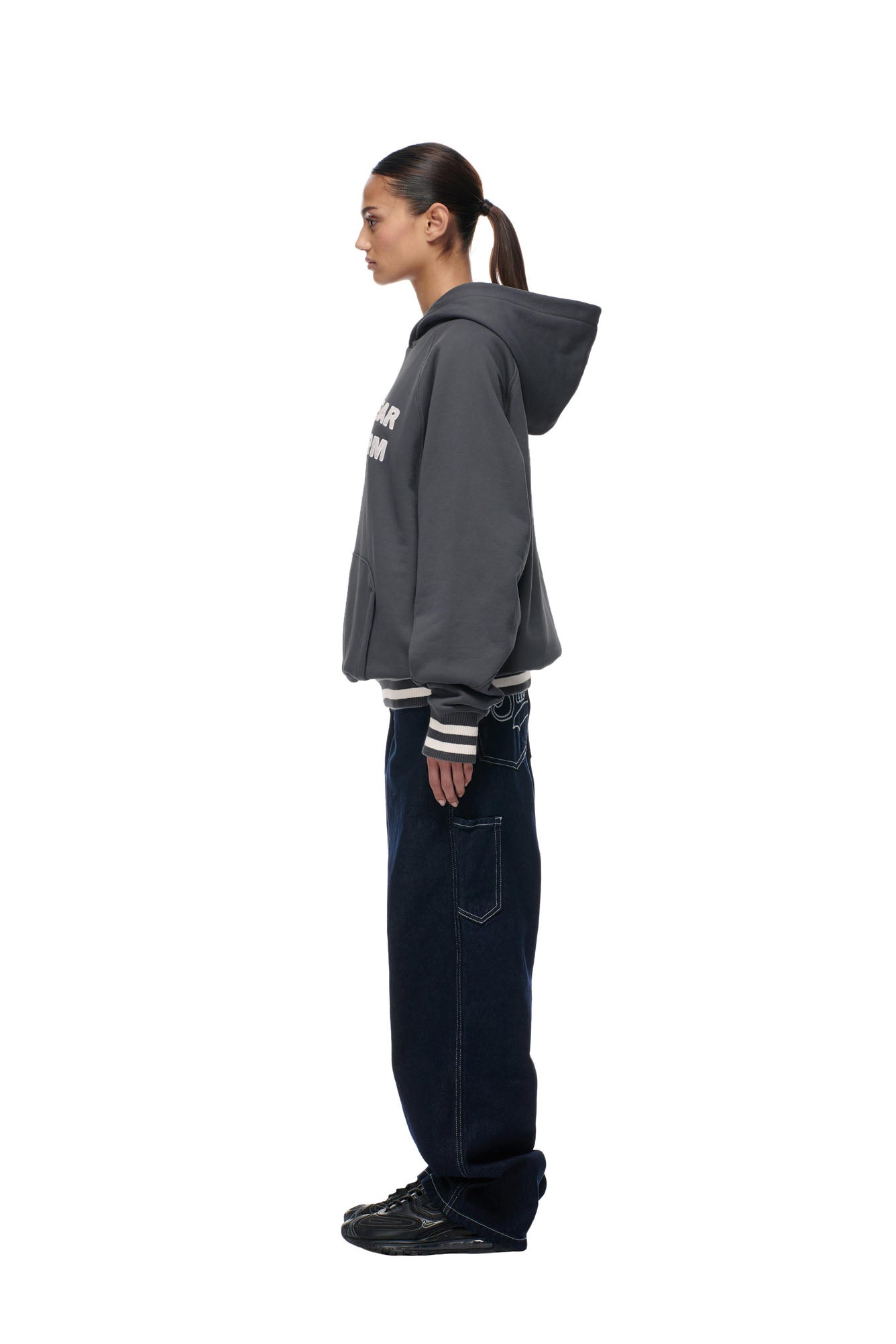 A woman with dark hair in a ponytail wears the 6PM ONLY WEAR REVERSIBLE HOODIE in anthracite/cream, along with dark baggy jeans and black sneakers, standing in profile against a plain white background.