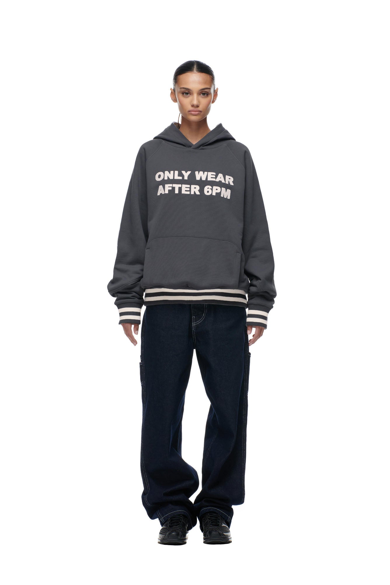 A person stands facing forward against a plain white background, wearing the 6PM ONLY WEAR REVERSIBLE HOODIE in anthracite/cream, dark loose jeans, and black sneakers.