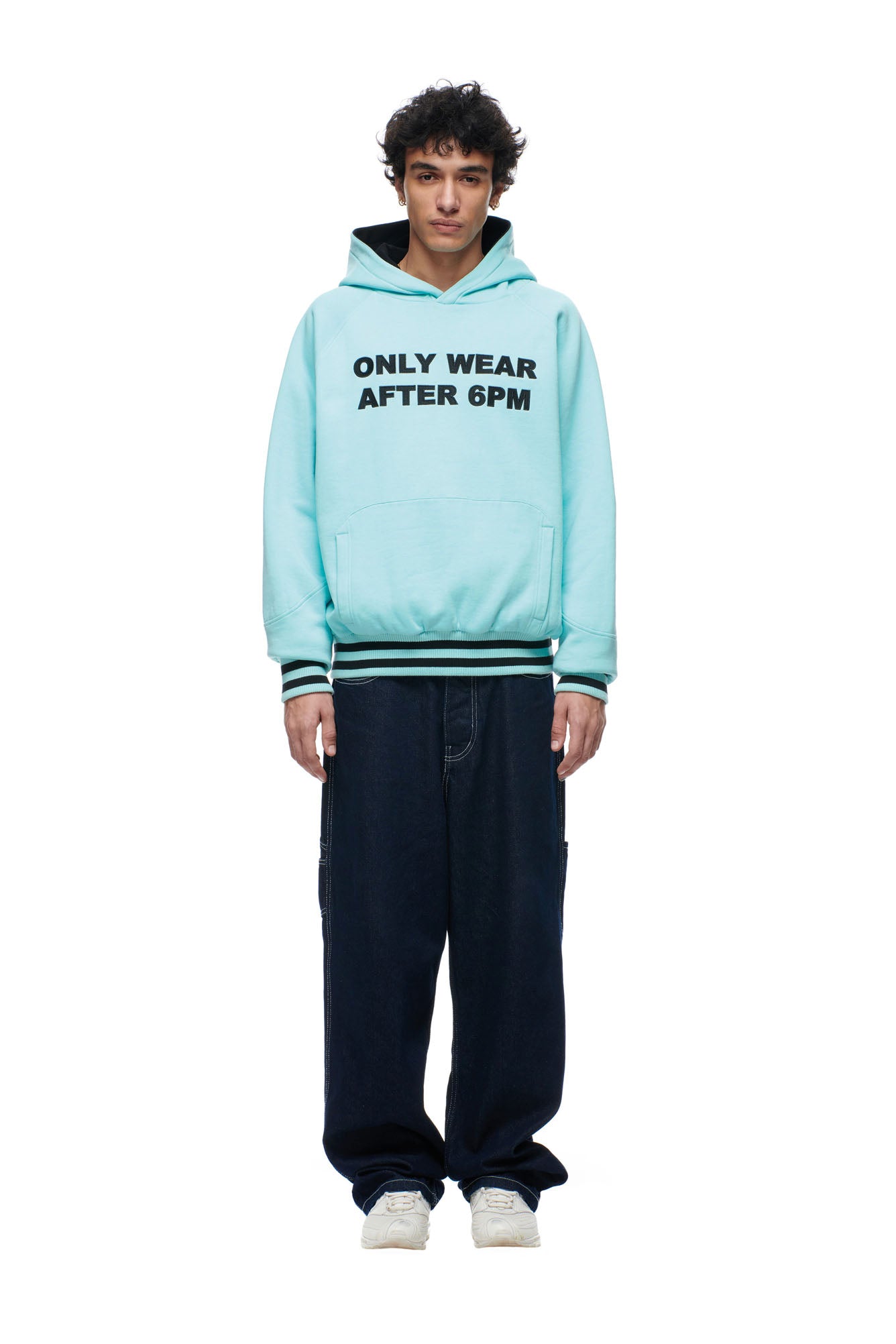 A young man faces forward in a 6PM ONLY WEAR REVERSIBLE HOODIE BLACK/TURQUOISE, dark baggy pants, and white sneakers, standing against a plain white background.