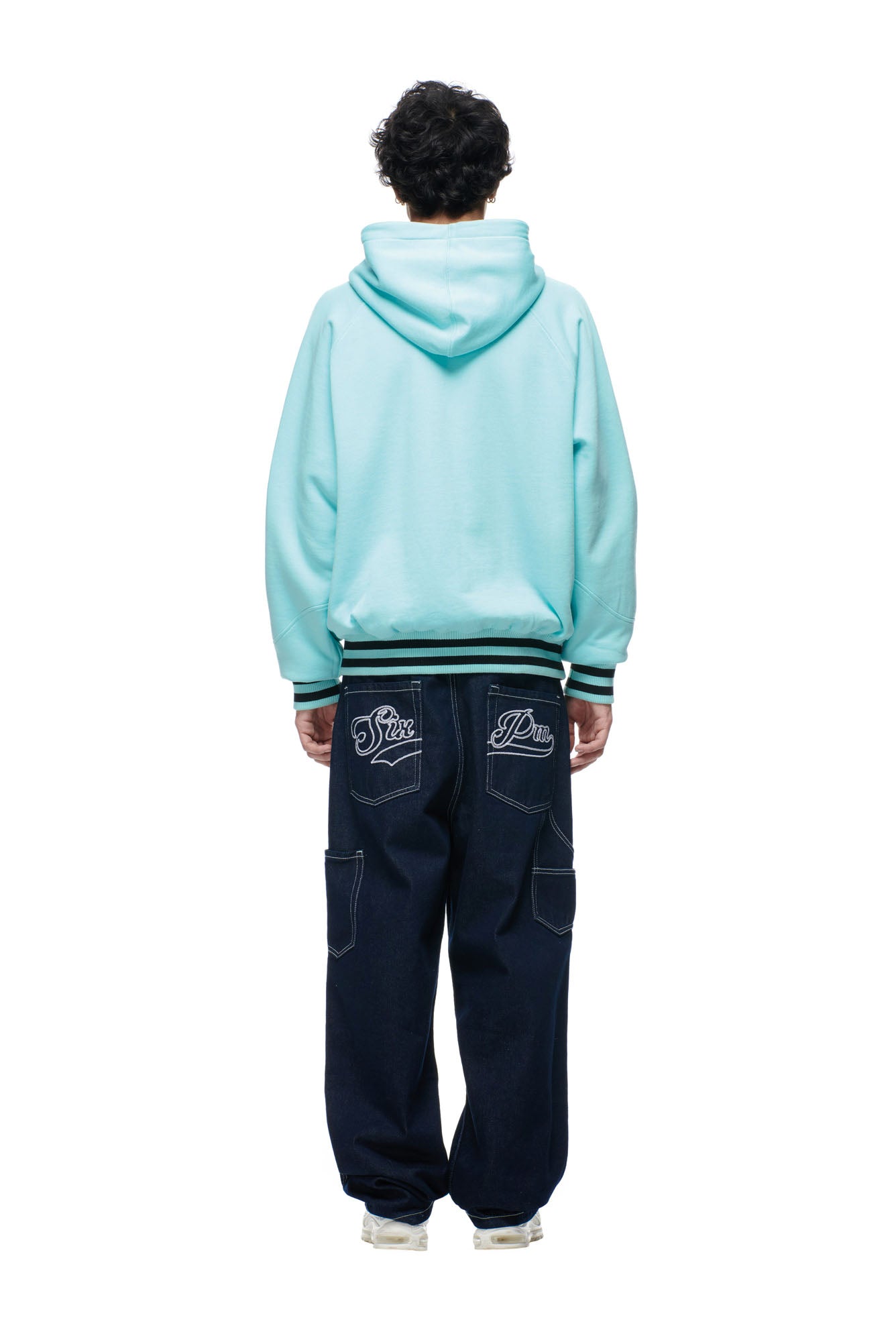 A person with short, curly hair wears the 6PM ONLY WEAR REVERSIBLE HOODIE in black/turquoise and loose dark blue jeans with white embroidery on the back pockets. They also sport white sneakers, standing against a plain white background.
