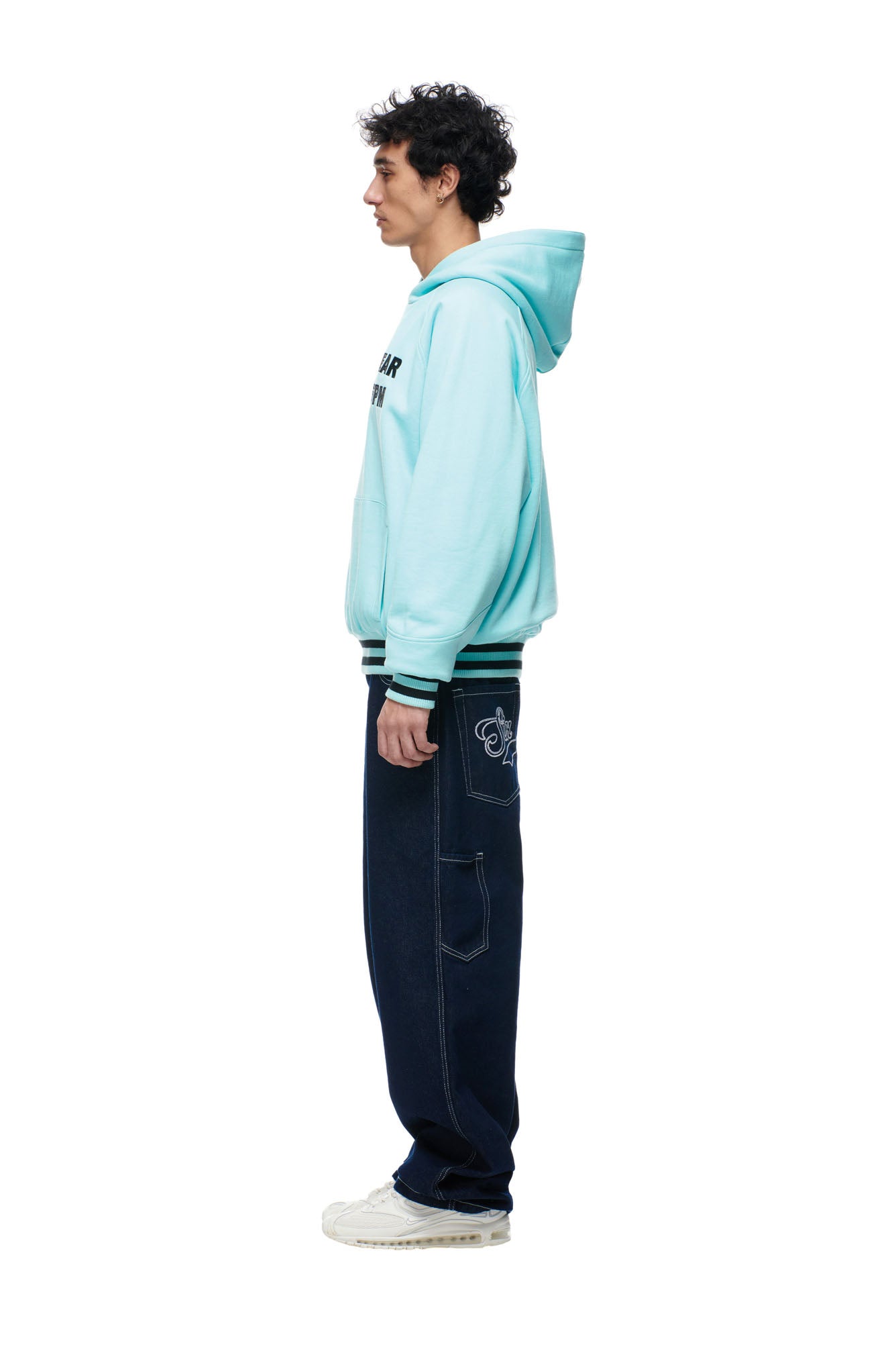 A man stands in profile against a white background, wearing the 6PM ONLY WEAR REVERSIBLE HOODIE BLACK/TURQUOISE, dark blue jeans with a patch on the side, and white sneakers.