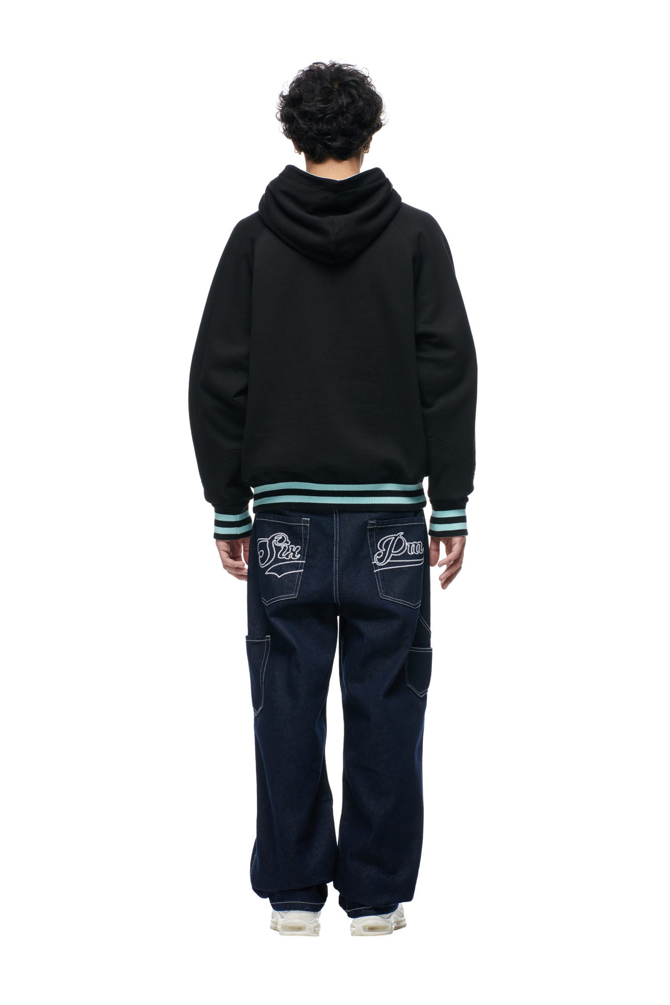 Wearing the 6PM ONLY WEAR REVERSIBLE HOODIE BLACK/TURQUOISE, a person with dark curly hair and white sneakers stands facing away, paired with loose dark blue jeans featuring embroidered back pockets against a plain white background.