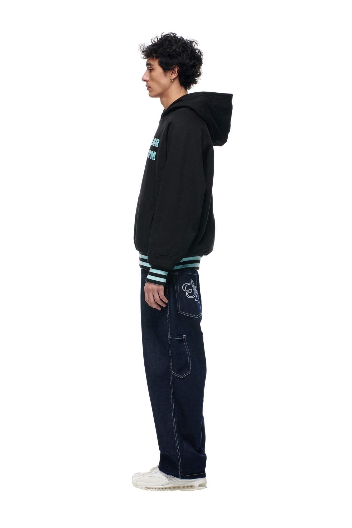 A person with curly dark hair stands in profile wearing the 6PM ONLY WEAR REVERSIBLE HOODIE in black/turquoise, paired with dark embroidered wide-leg jeans and white sneakers, set against a plain white background.
