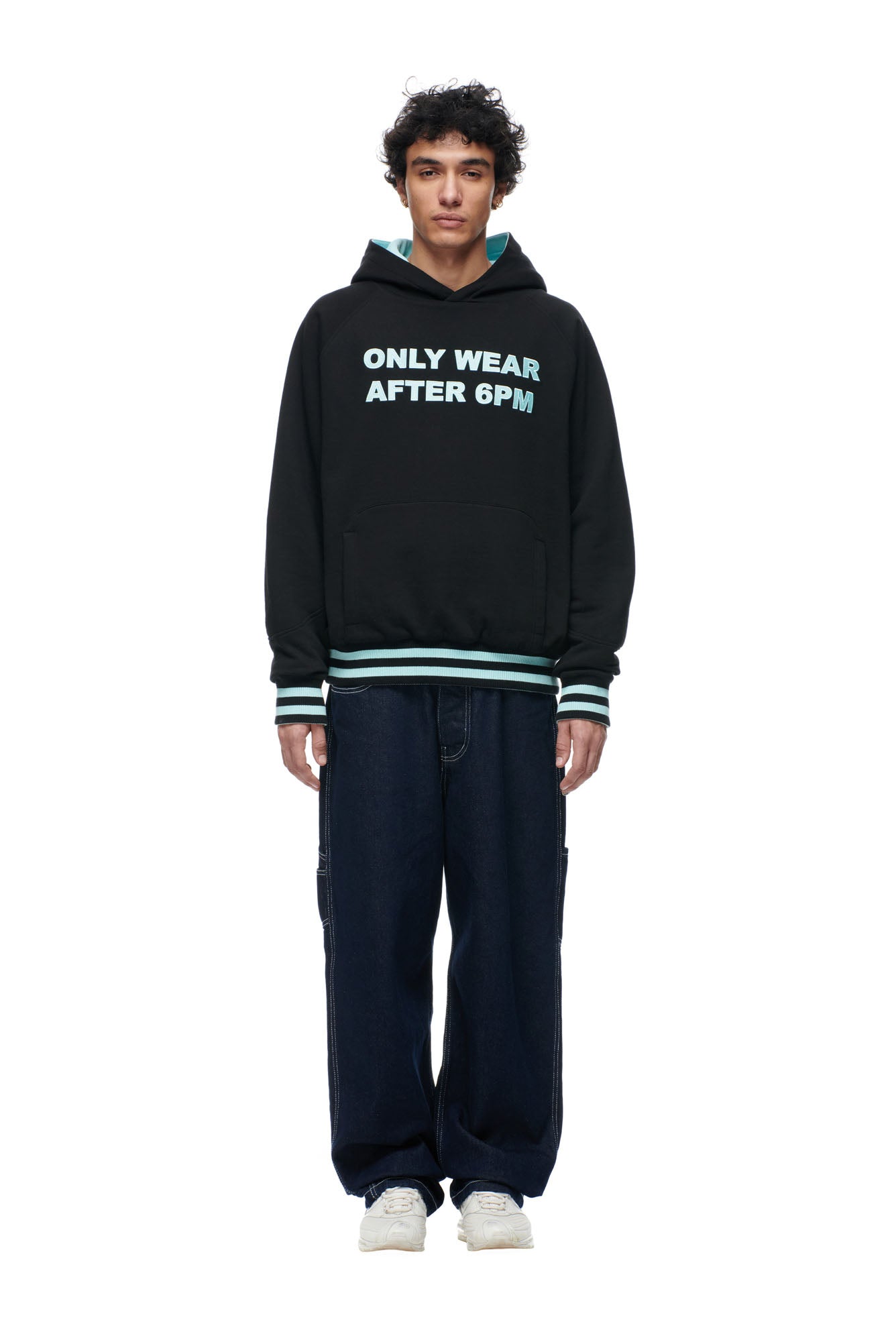A person faces forward wearing the 6PM ONLY WEAR REVERSIBLE HOODIE BLACK/TURQUOISE with "ONLY WEAR AFTER 6PM" text, striped cuffs and hem, dark loose jeans, and white sneakers against a plain white background.