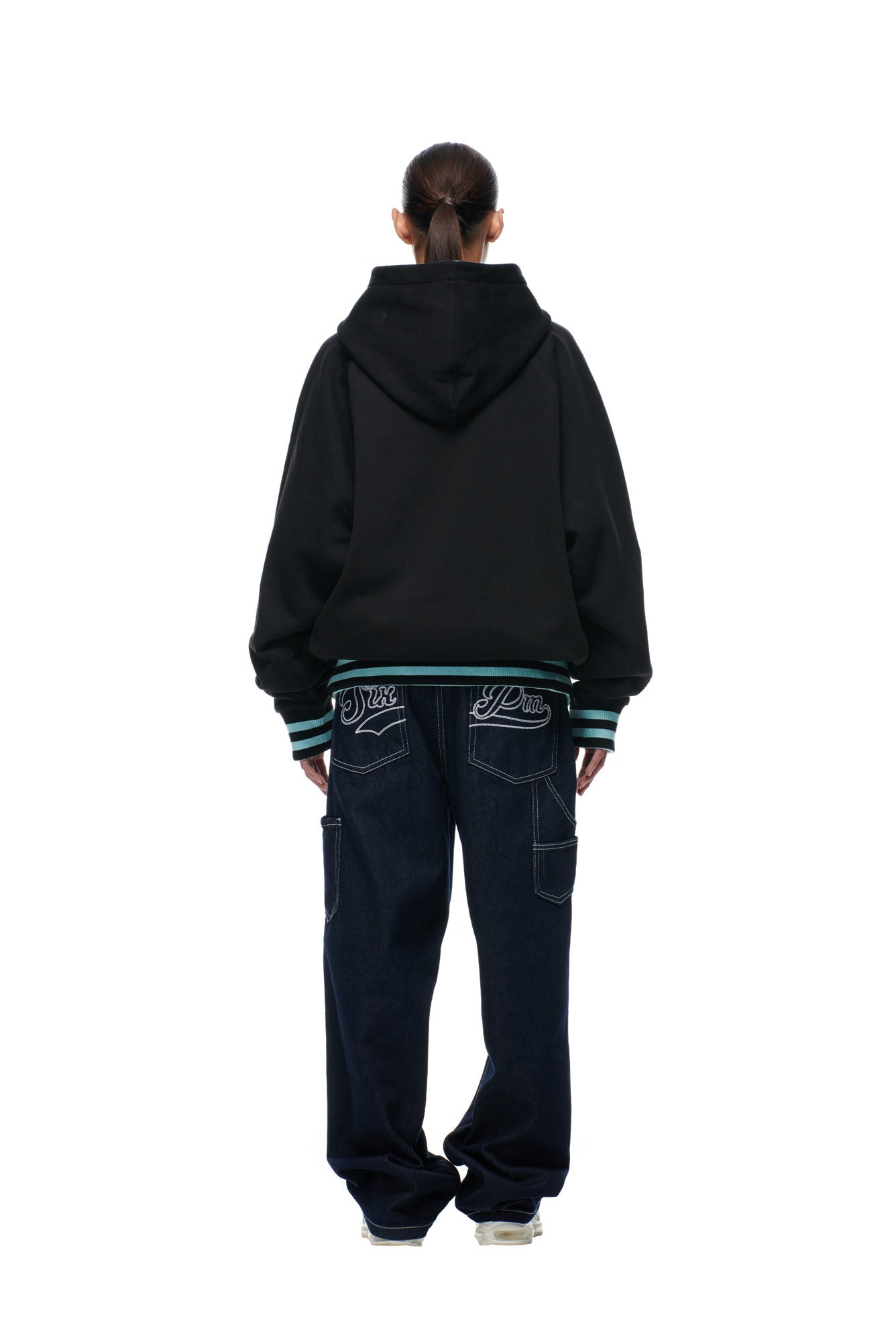 A person stands facing away against a white background, wearing the 6PM ONLY WEAR REVERSIBLE HOODIE in black/turquoise, dark baggy jeans with white-embroidered back pockets, white sneakers, and their hair tied back.