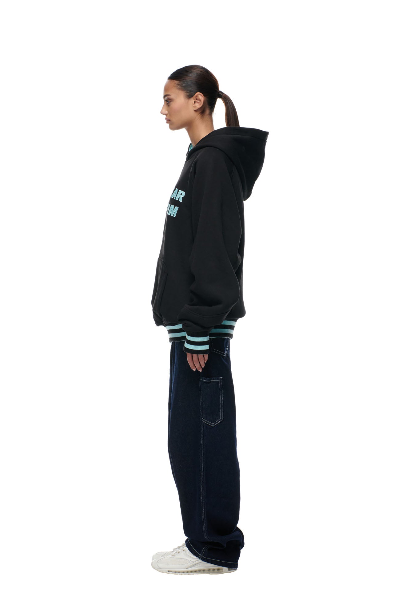 A person stands in profile against a white background, wearing the 6PM ONLY WEAR REVERSIBLE HOODIE BLACK/TURQUOISE, dark loose jeans, and white sneakers. Their hair is tied back in a low ponytail.