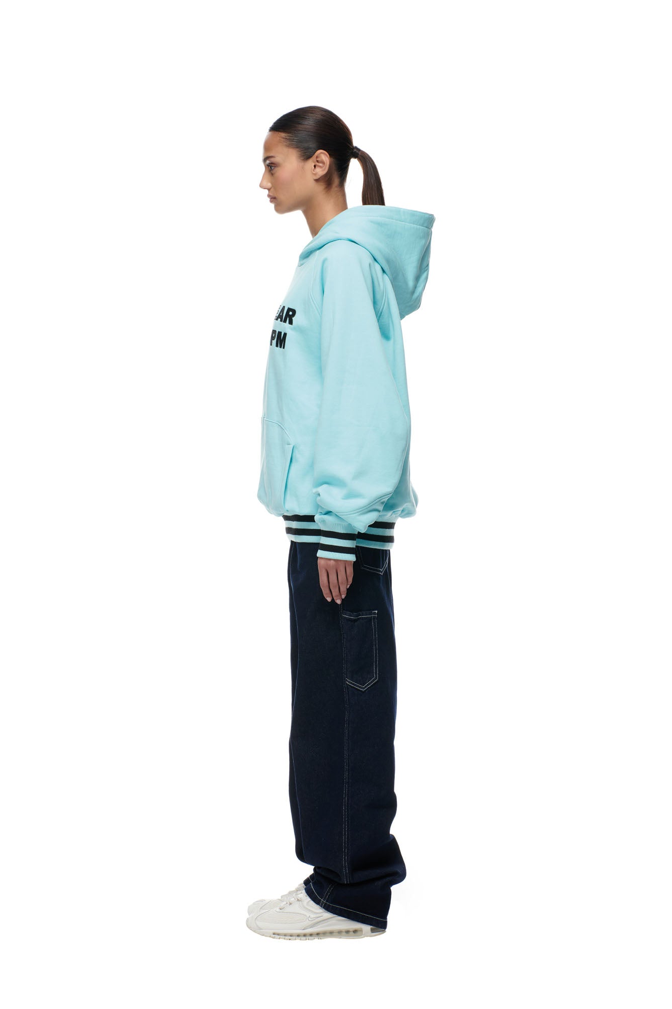 A person with dark hair in a ponytail stands in profile, wearing the 6PM ONLY WEAR REVERSIBLE HOODIE BLACK/TURQUOISE, dark jeans, and white sneakers, against a plain white background.