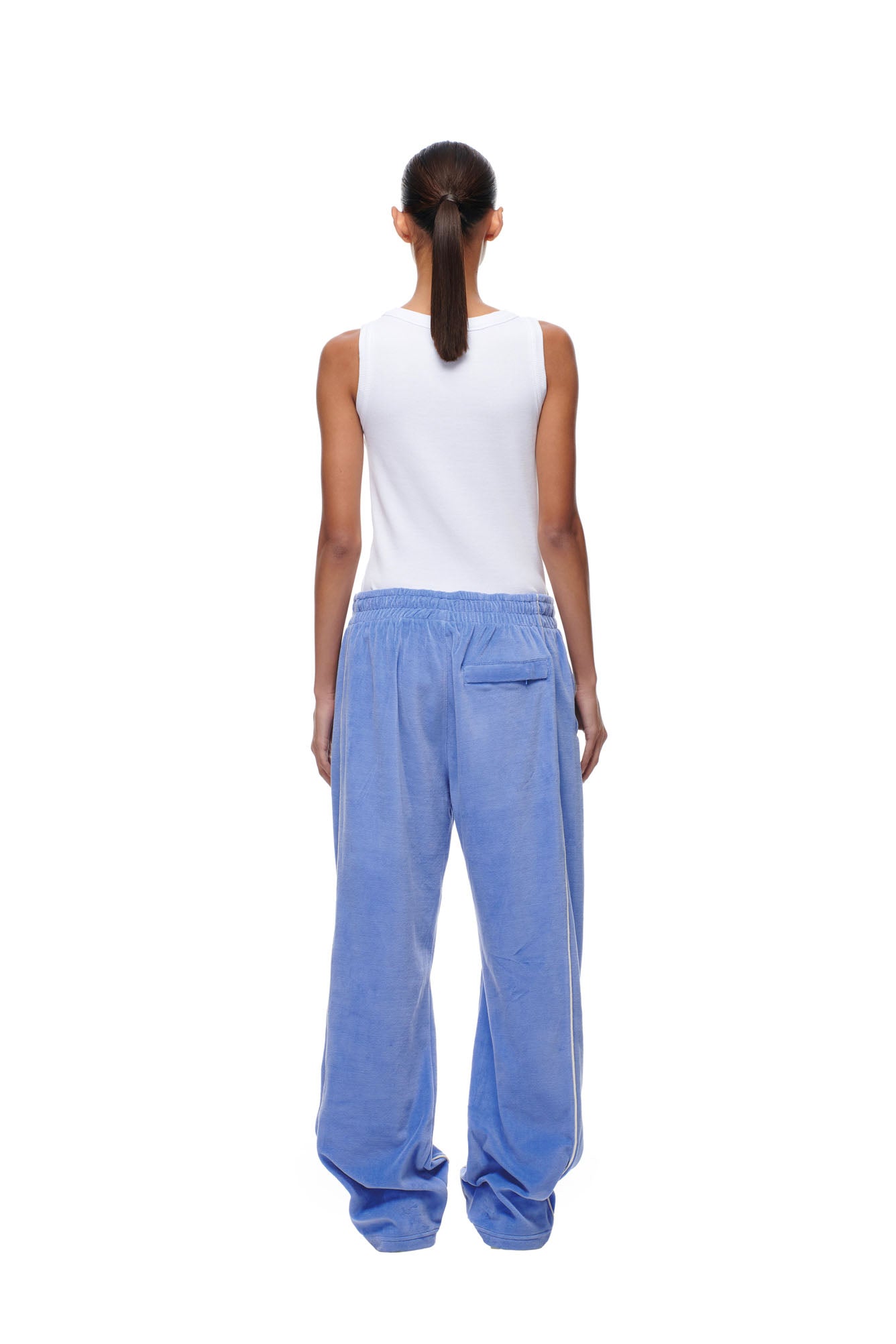 A person with a ponytail stands facing away against a white background, wearing a white sleeveless top and 6PM BF Velours Trackpants in Persian Blue featuring a back pocket.