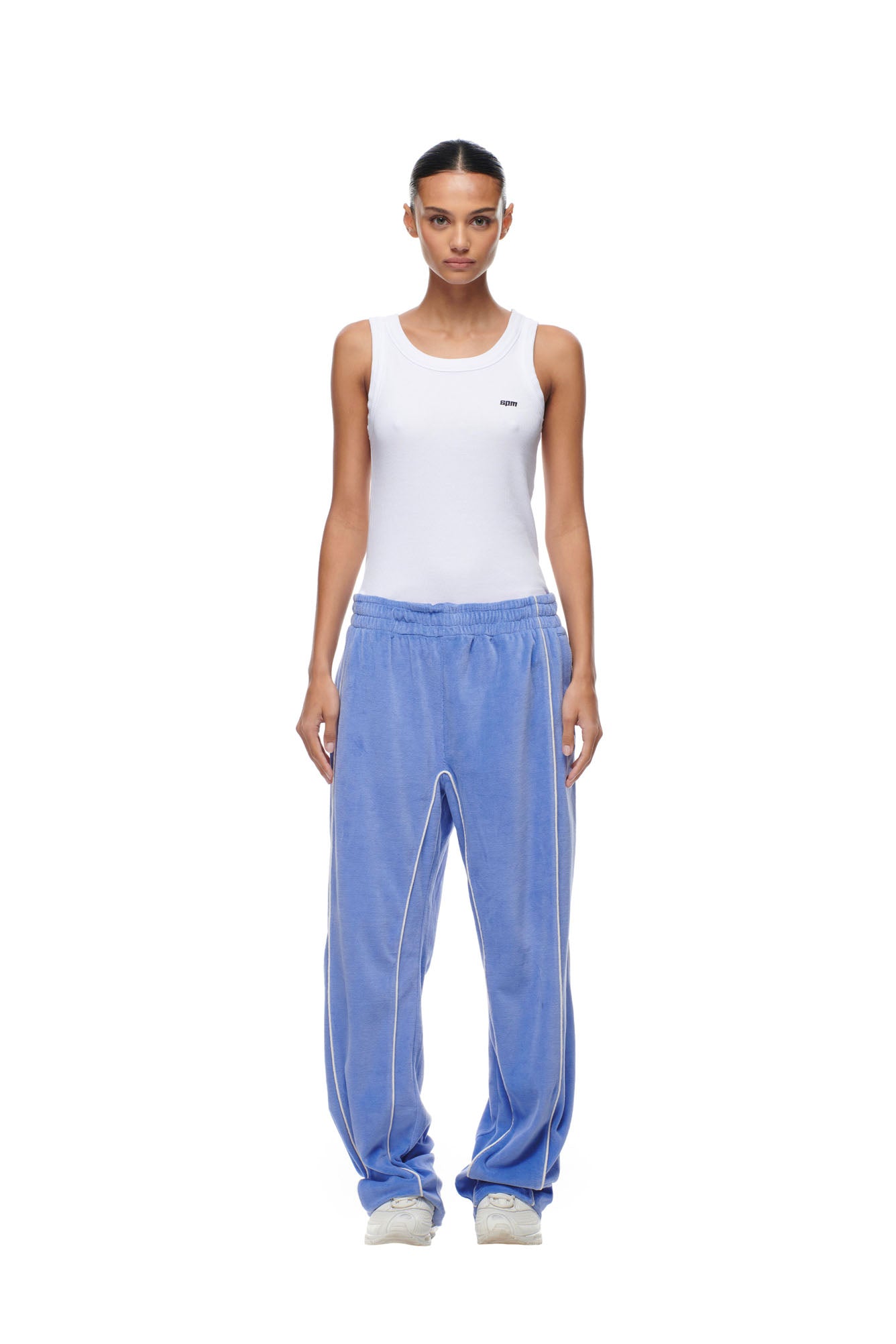 A woman stands facing forward on a plain white background, wearing 6PM BF VELOURS TRACKPANTS in Persian Blue with white piping, a white sleeveless tank top featuring a small logo, and white sneakers.