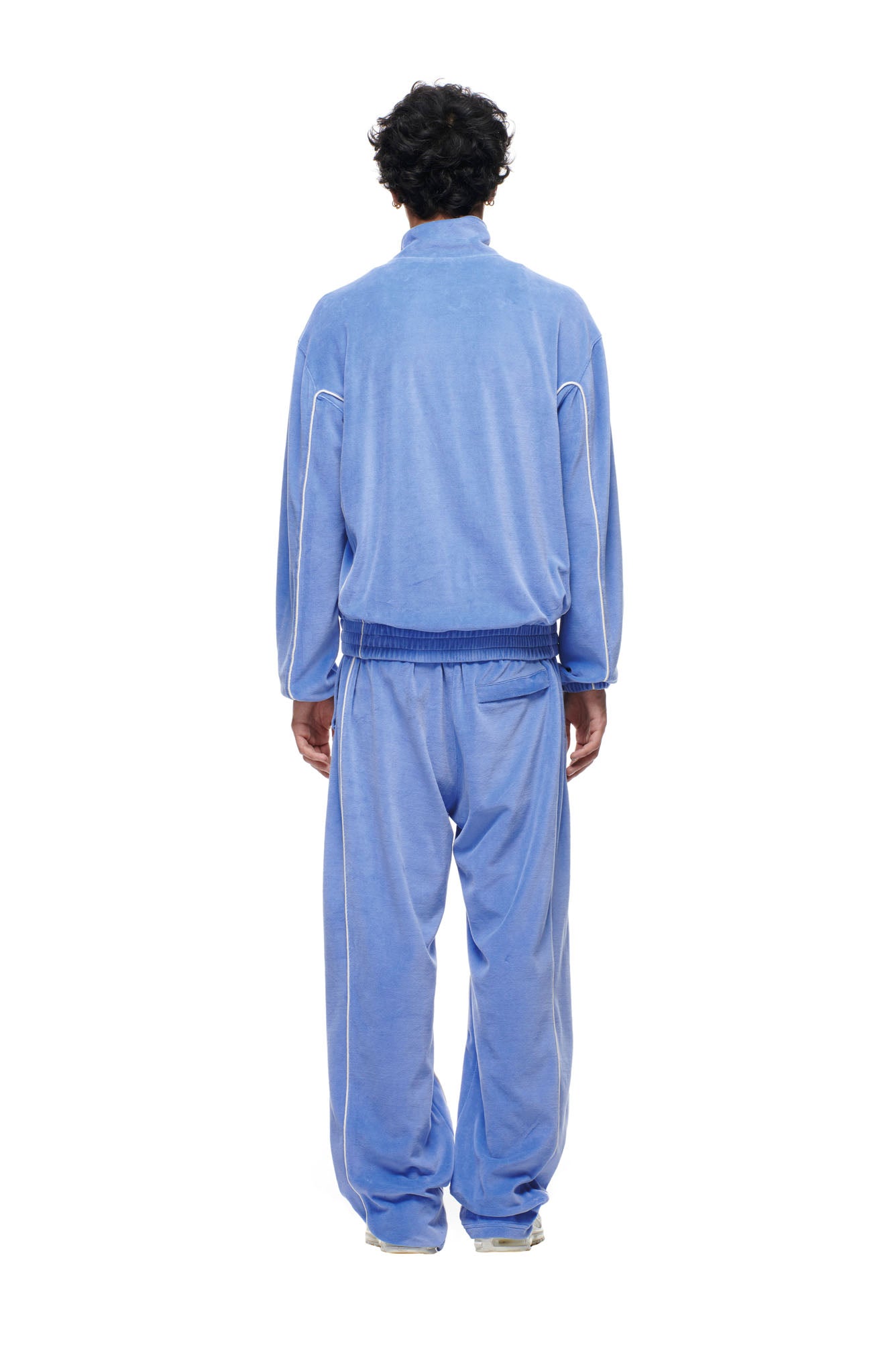 A person with short curly hair, seen from behind, wears the 6PM BF VELOURS TRACKJACKET in Persian Blue, featuring a loose fit and white piping, standing against a plain white background.