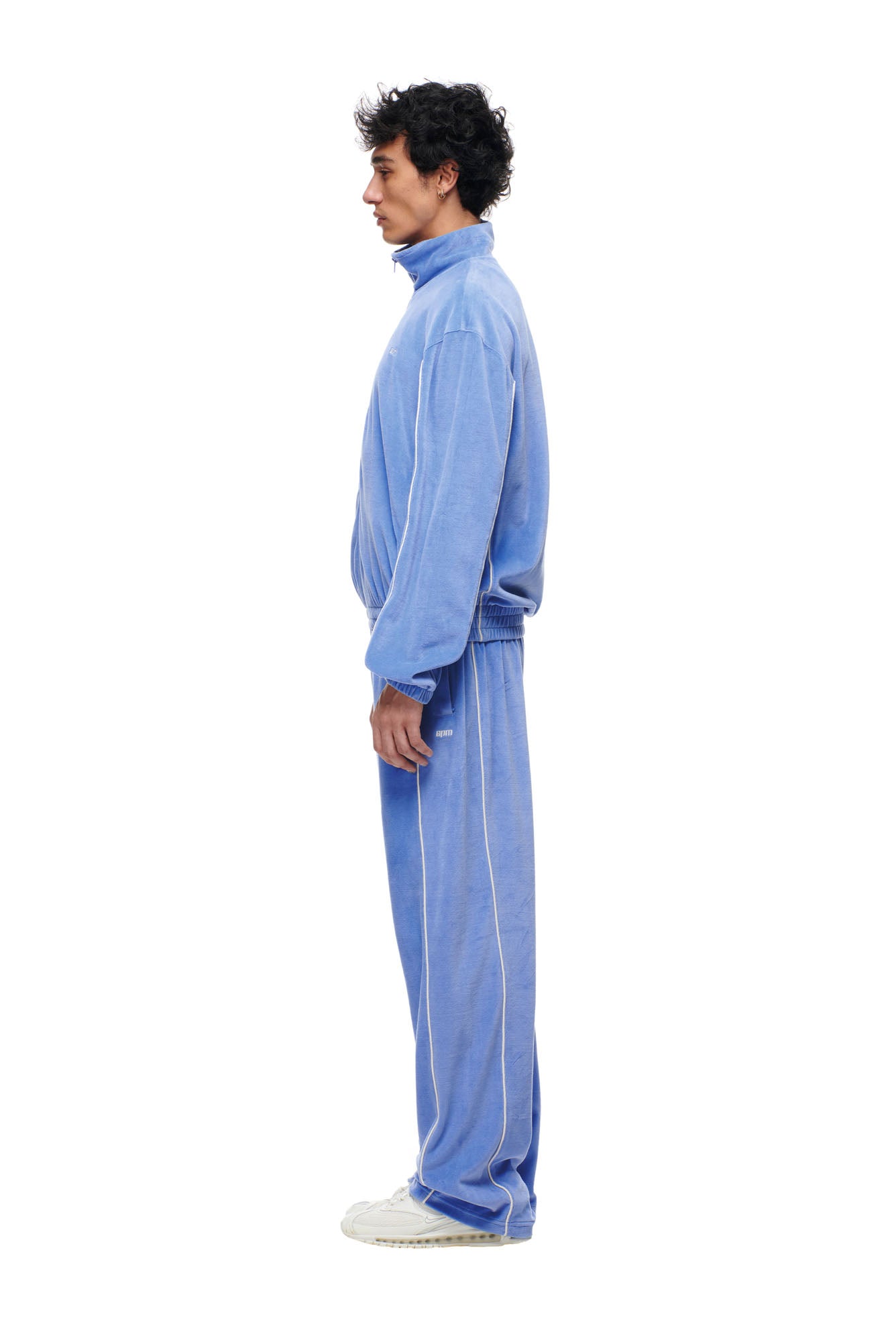 A man stands in profile, facing left, wearing the 6PM BF Velours Trackjacket in Persian Blue and white sneakers against a plain white background.