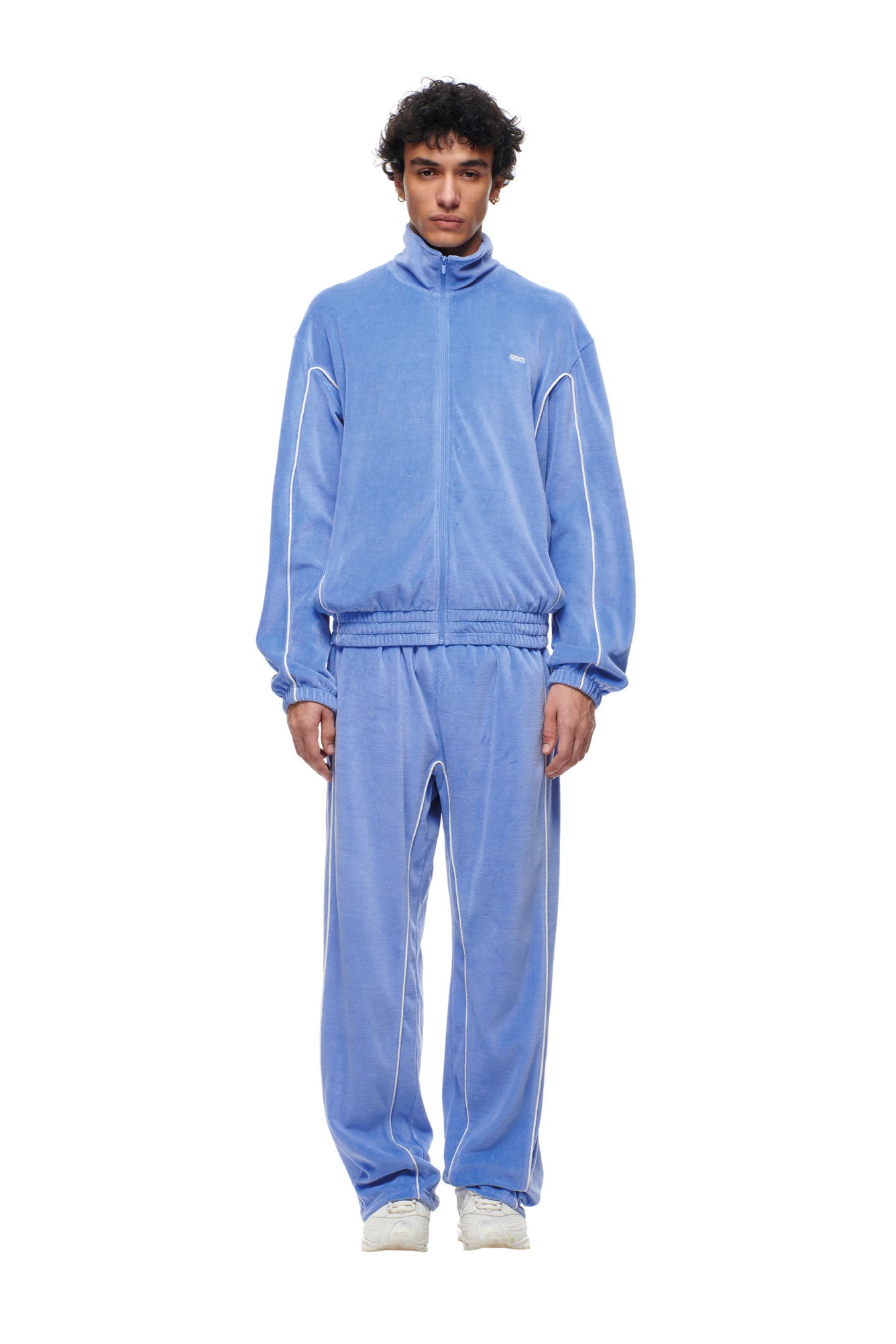 A man with curly hair faces forward, wearing the 6PM BF VELOURS TRACKJACKET in Persian Blue, paired with white sneakers and a neutral expression, set against a plain white background.