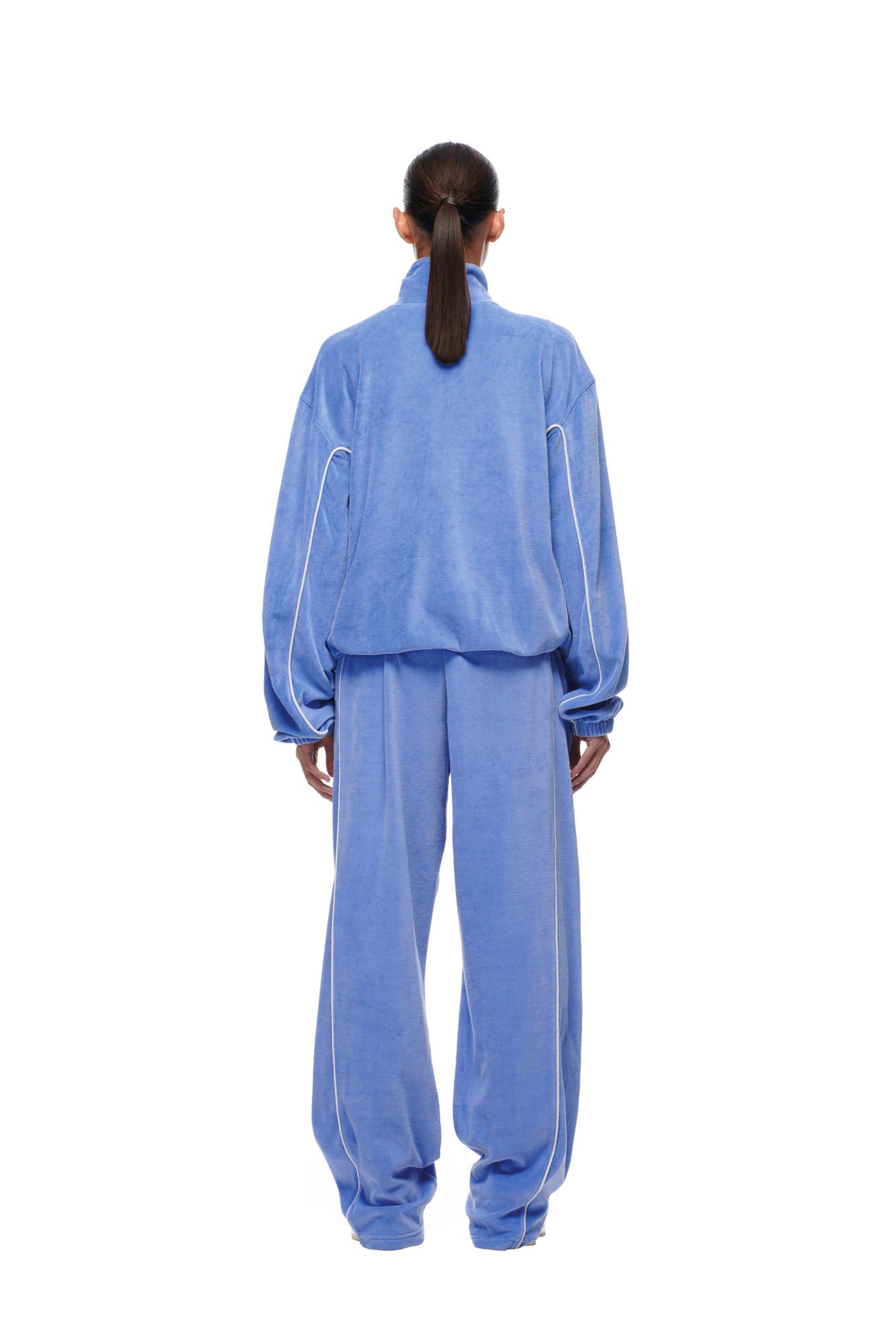 A person with long dark hair in a low ponytail stands facing away, wearing the 6PM BF VELOURS TRACKJACKET PERSIAN BLUE, a loose-fitting light blue velour jacket with white piping, against a plain white background.