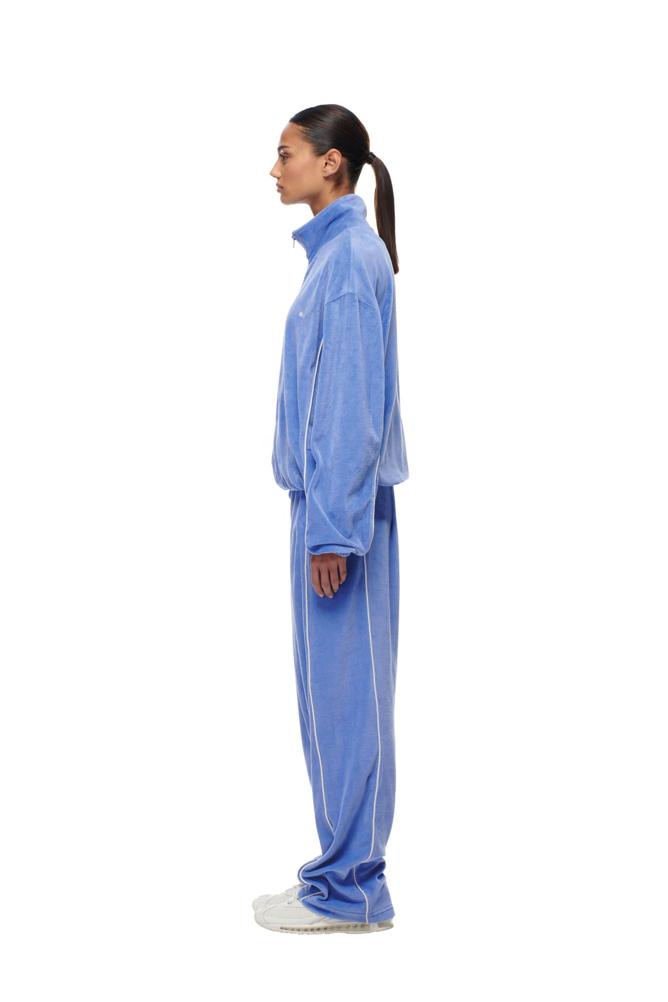 A woman in profile wears the 6PM BF VELOURS TRACKJACKET in Persian Blue with white piping, paired with white sneakers and her hair tied back, standing against a plain white background.