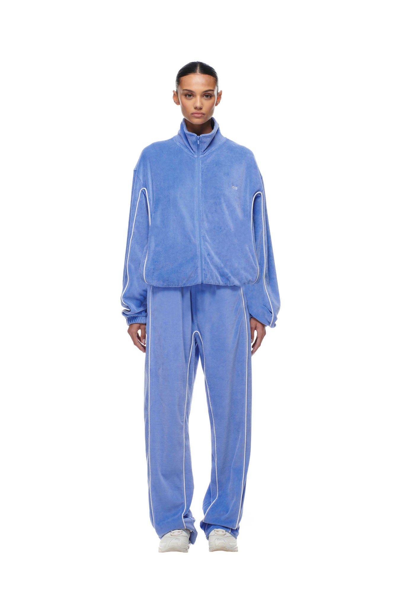A person stands facing forward against a white background, wearing the 6PM BF VELOURS TRACKJACKET in Persian Blue with white piping and matching wide-leg pants, paired with white sneakers.