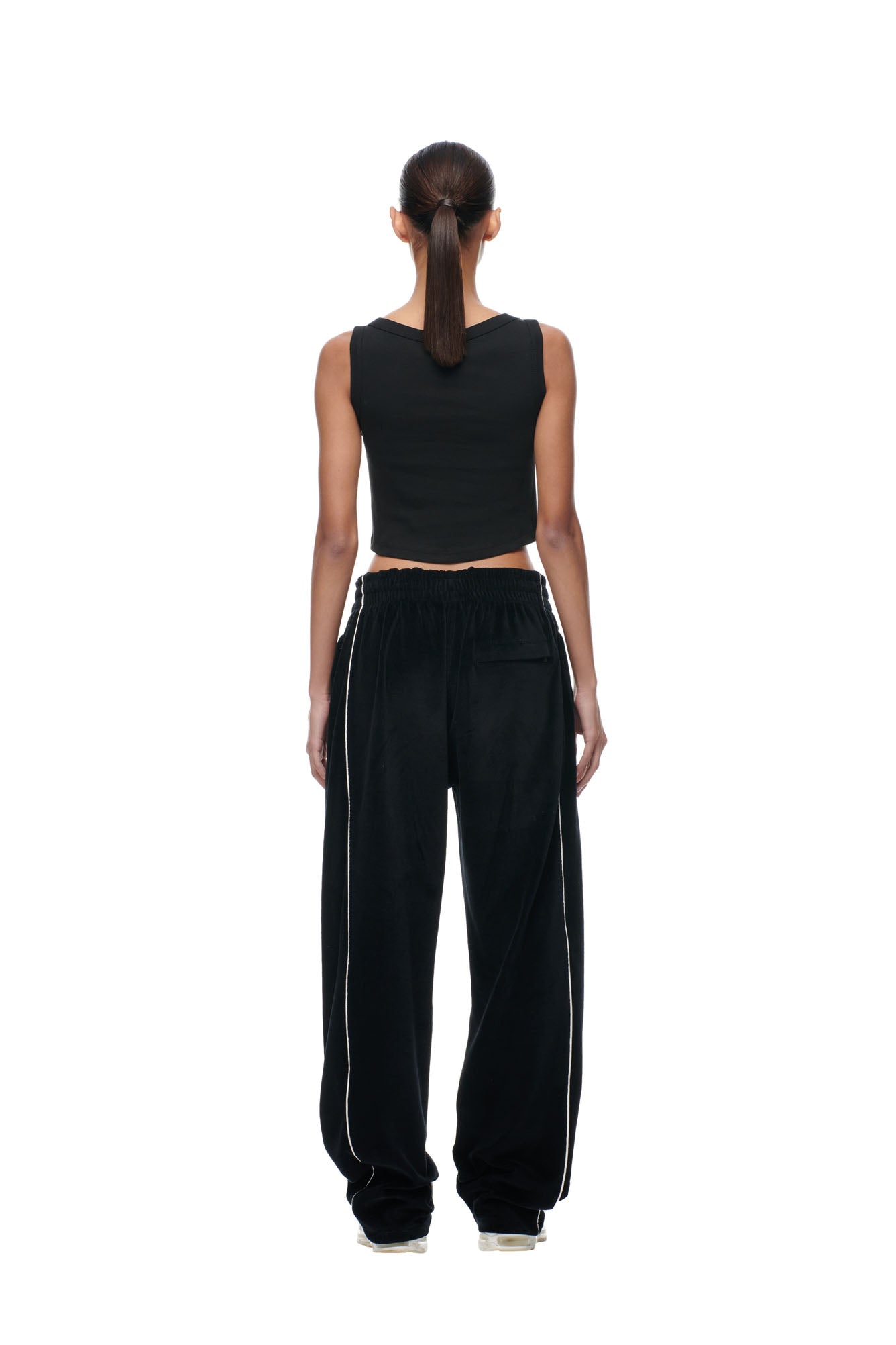A person with a long ponytail stands facing away against a plain white background, wearing a black sleeveless crop top and 6PM's BF VELOURS TRACKPANTS BLACK featuring white piping.