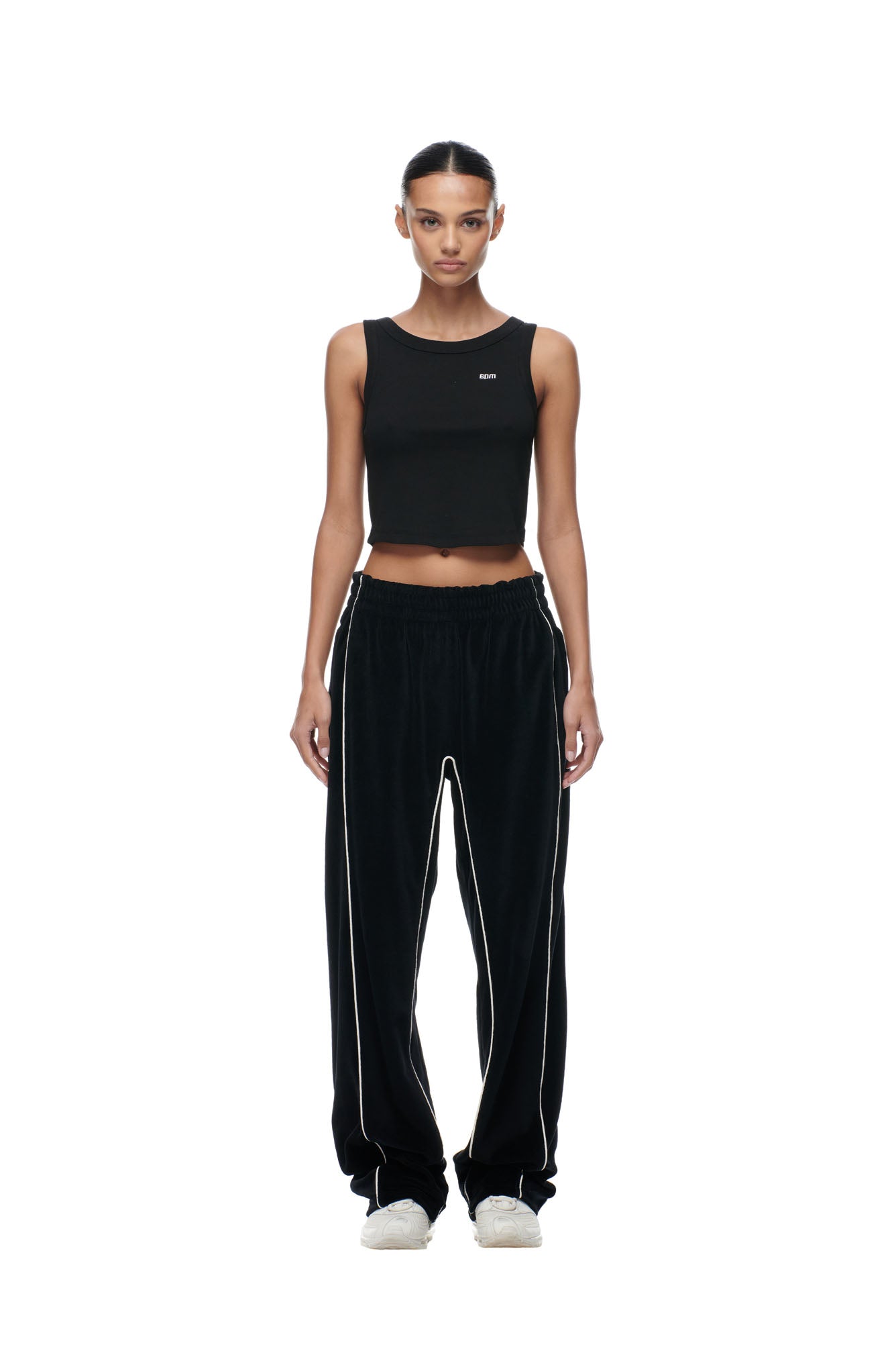 A woman stands facing forward, wearing 6PM BF VELOURS TRACKPANTS BLACK, a black sleeveless crop top, and white sneakers against a plain white background.