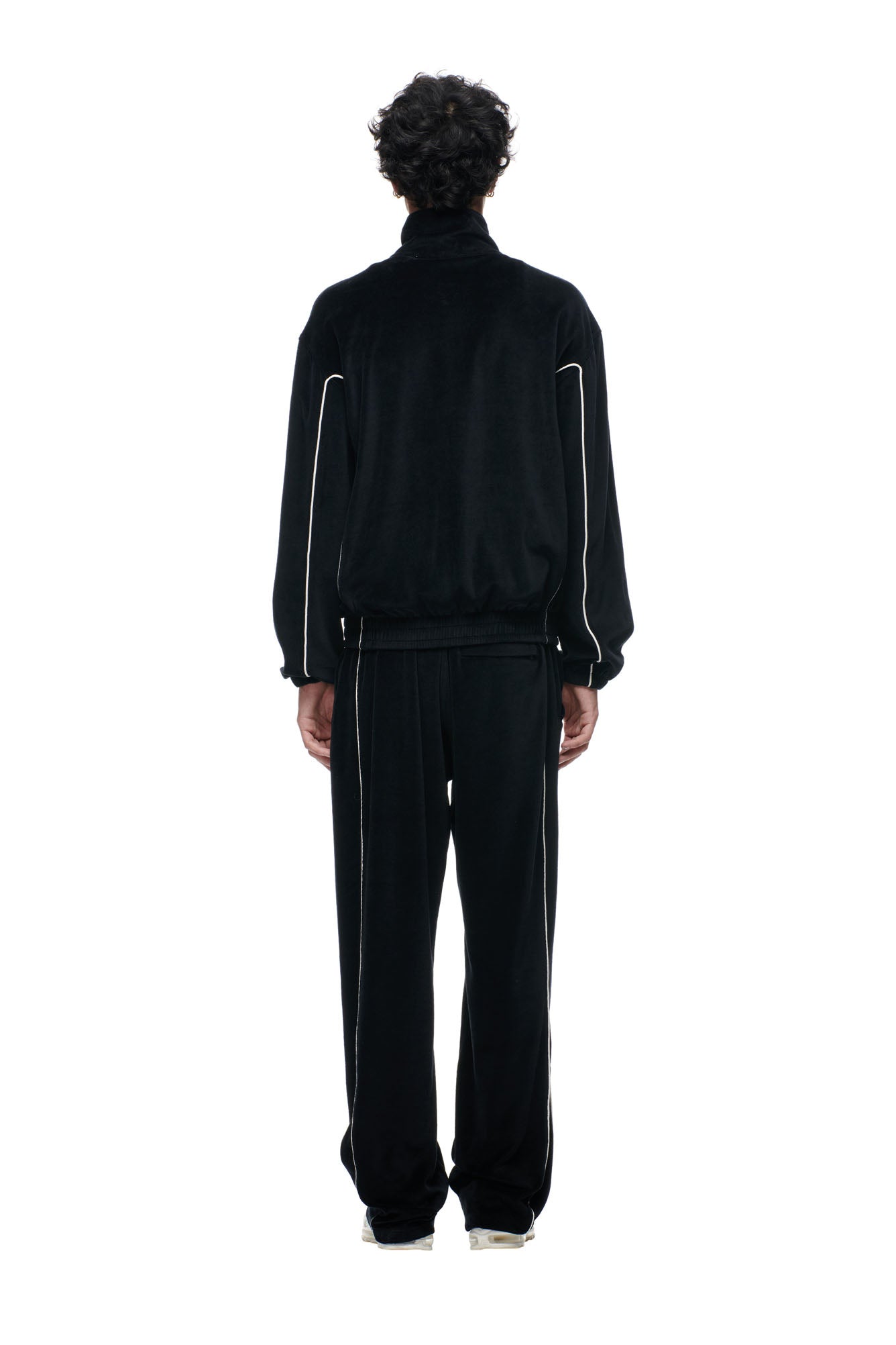 A person stands facing away from the camera, wearing the 6PM BF VELOURS TRACKJACKET BLACK with white piping on the sleeves and pants, paired with white sneakers, set against a plain white background.