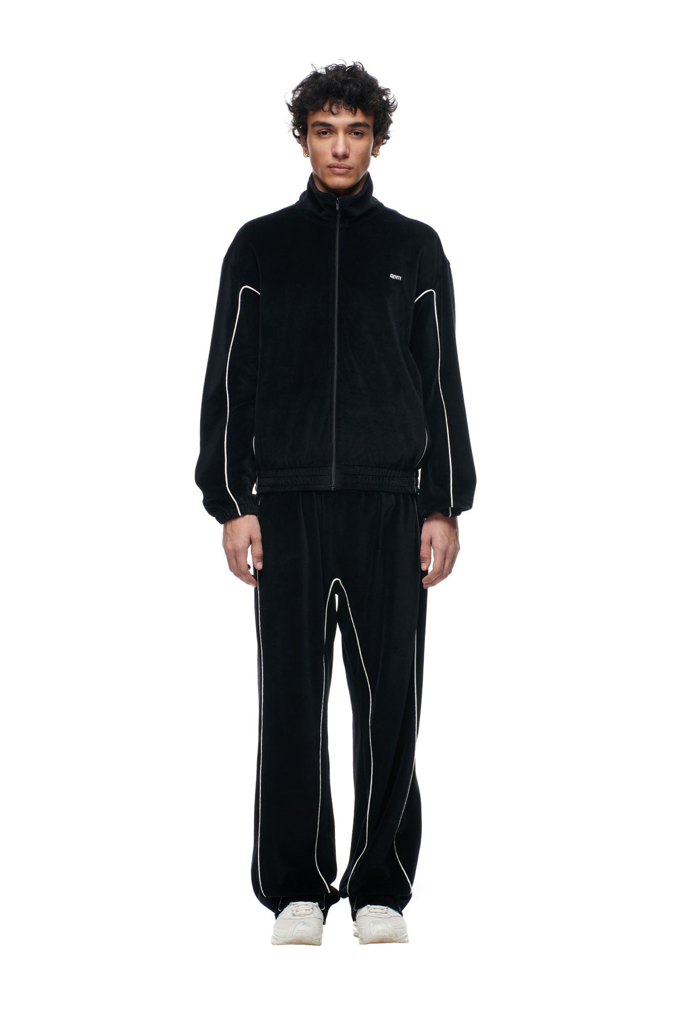 A person faces forward against a white background, wearing the 6PM BF VELOURS TRACKJACKET BLACK with matching black track pants featuring white piping, and white sneakers.