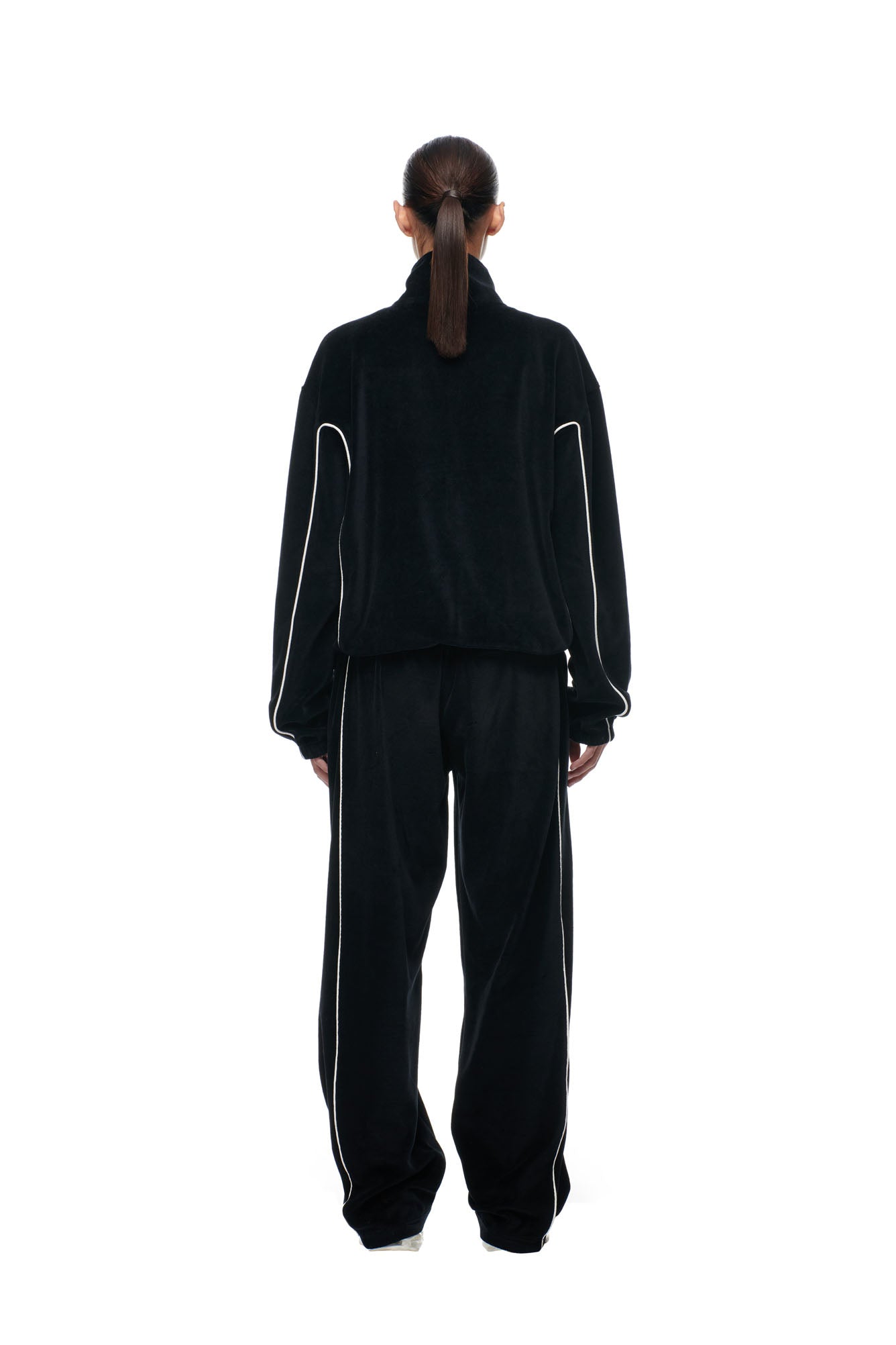 A person stands with their back to the camera, wearing the 6PM BF VELOURS TRACKJACKET BLACK, featuring white piping. They pair it with white shoes and have their hair in a low ponytail, all set against a plain white background.