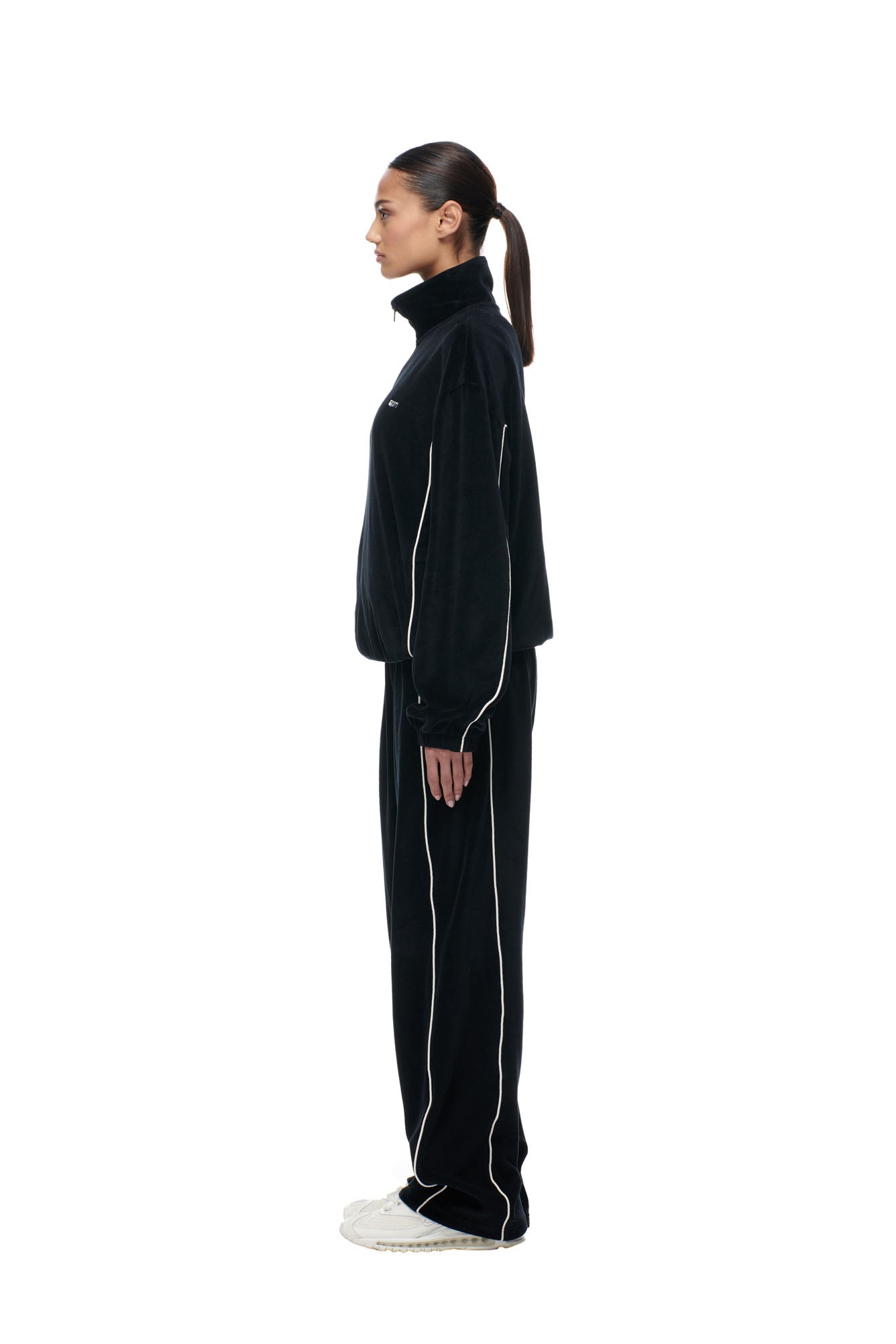 A woman stands in profile against a white background, wearing the 6PM BF VELOURS TRACKJACKET BLACK with white piping and matching pants, paired with white sneakers. Her hair is tied back in a low ponytail.