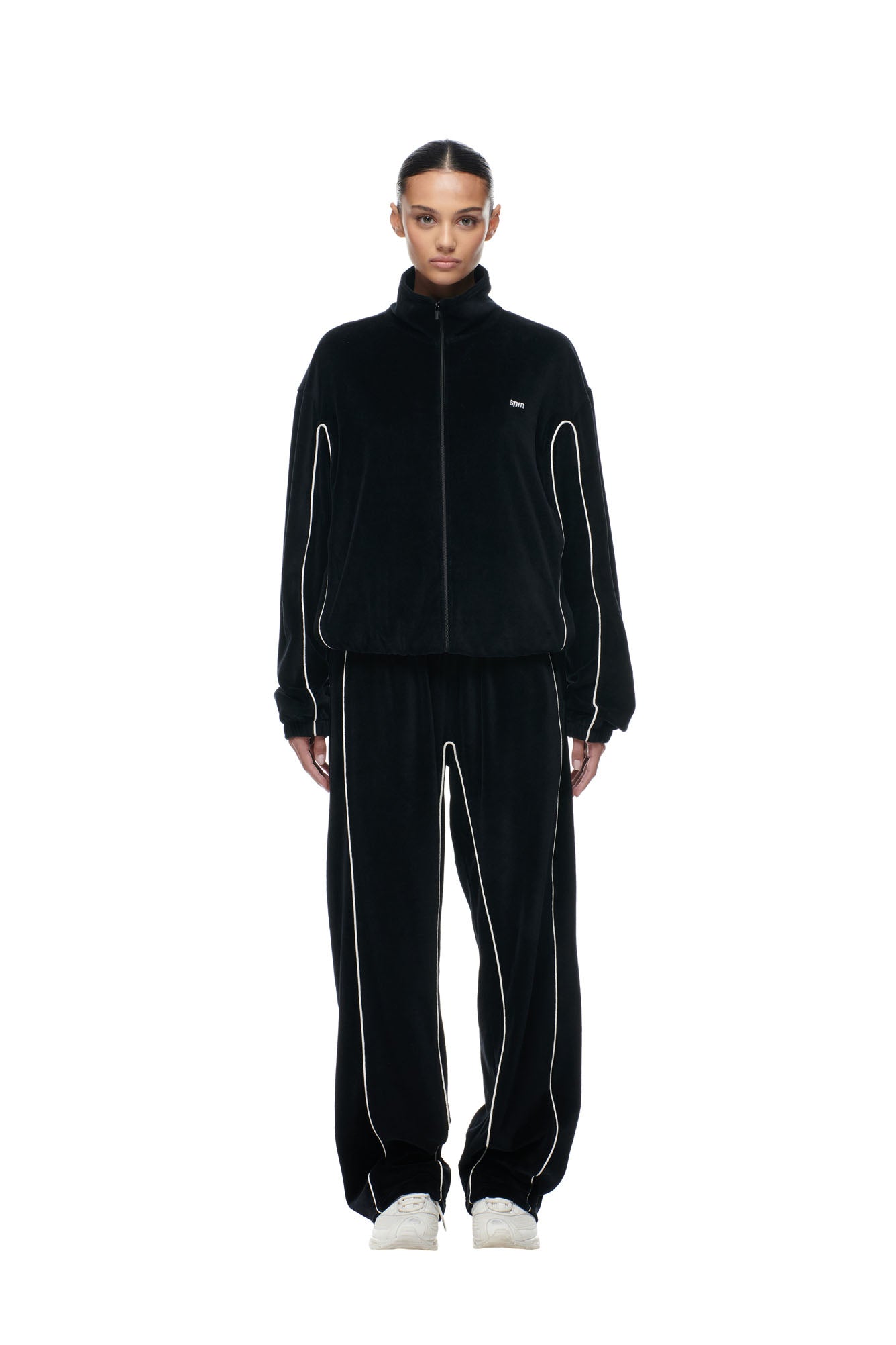A person stands facing forward in the 6PM BF VELOURS TRACKJACKET BLACK, styled with white piping on the sleeves and paired with matching pants and white sneakers, set against a plain white background.