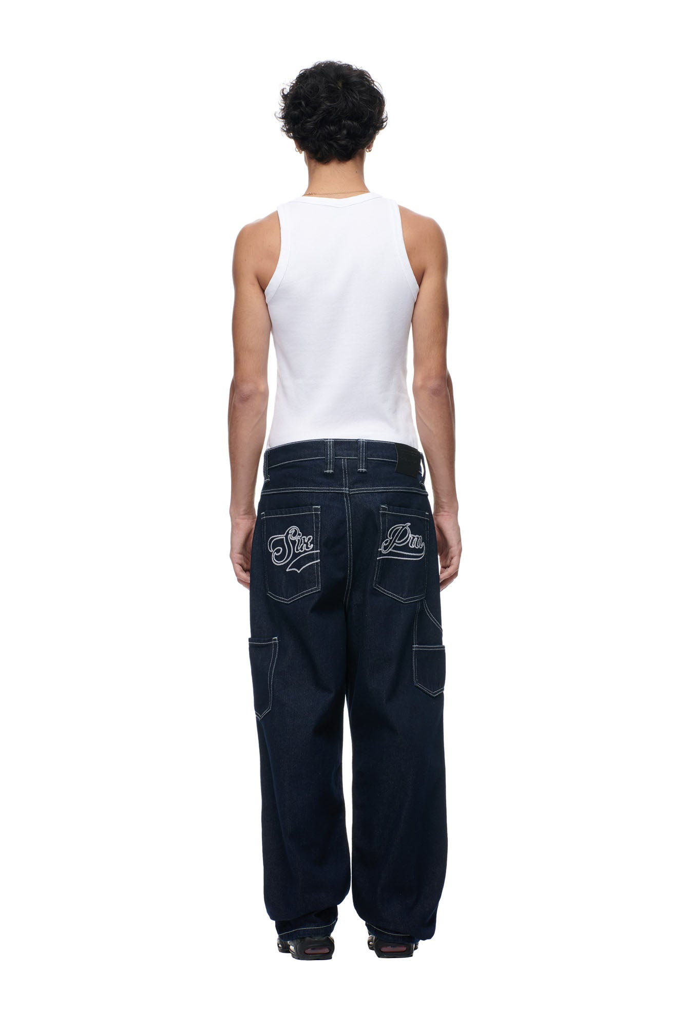 A person with short, curly hair is seen from behind, wearing a white tank and 6PM's SCRIPT LOGO DENIM—loose dark blue jeans featuring white embroidered back pocket designs—against a plain white background.