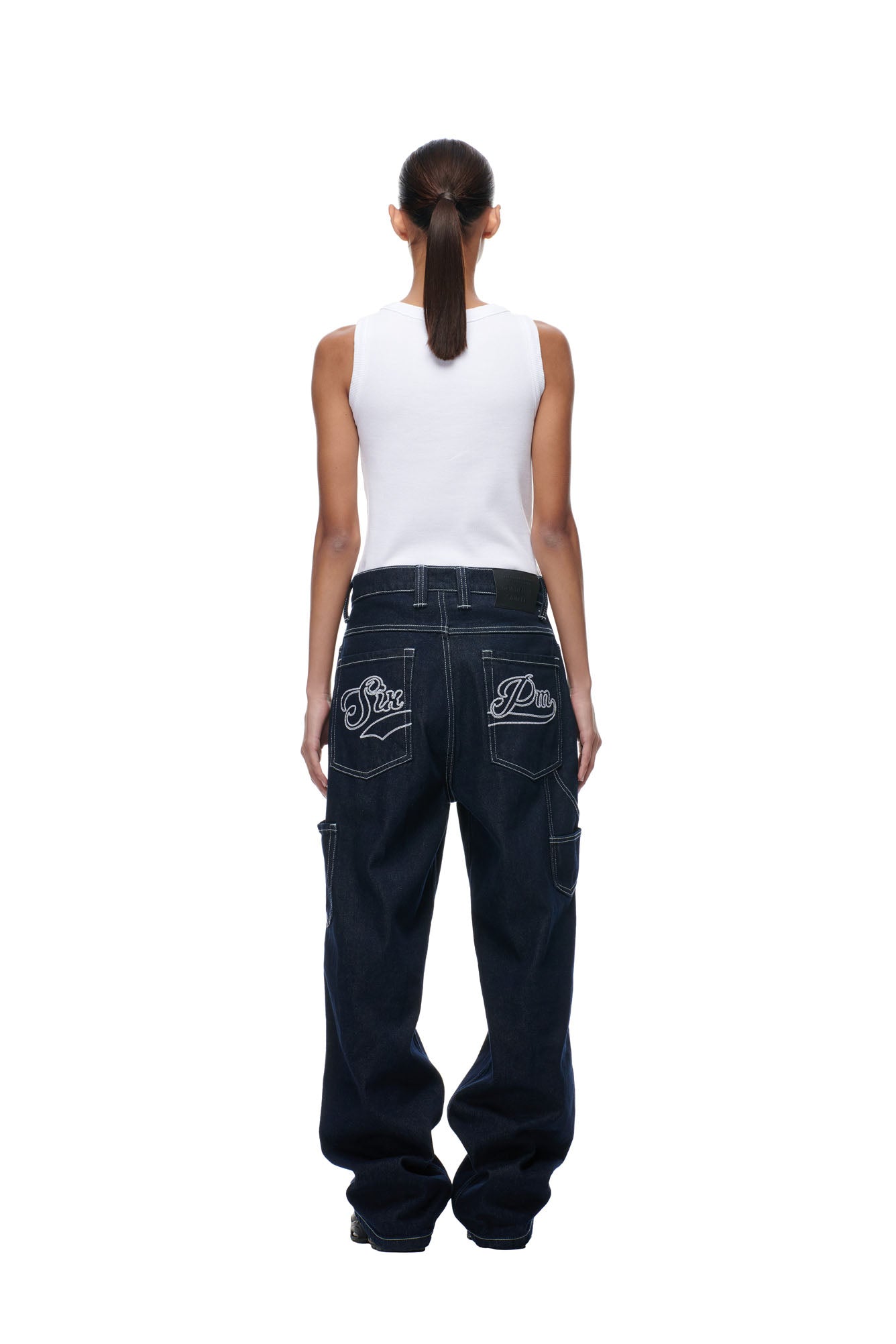 A person with long dark hair in a ponytail wears a white sleeveless top and 6PM SCRIPT LOGO DENIM, featuring baggy dark jeans with embroidered back pockets, standing facing away against a white background.