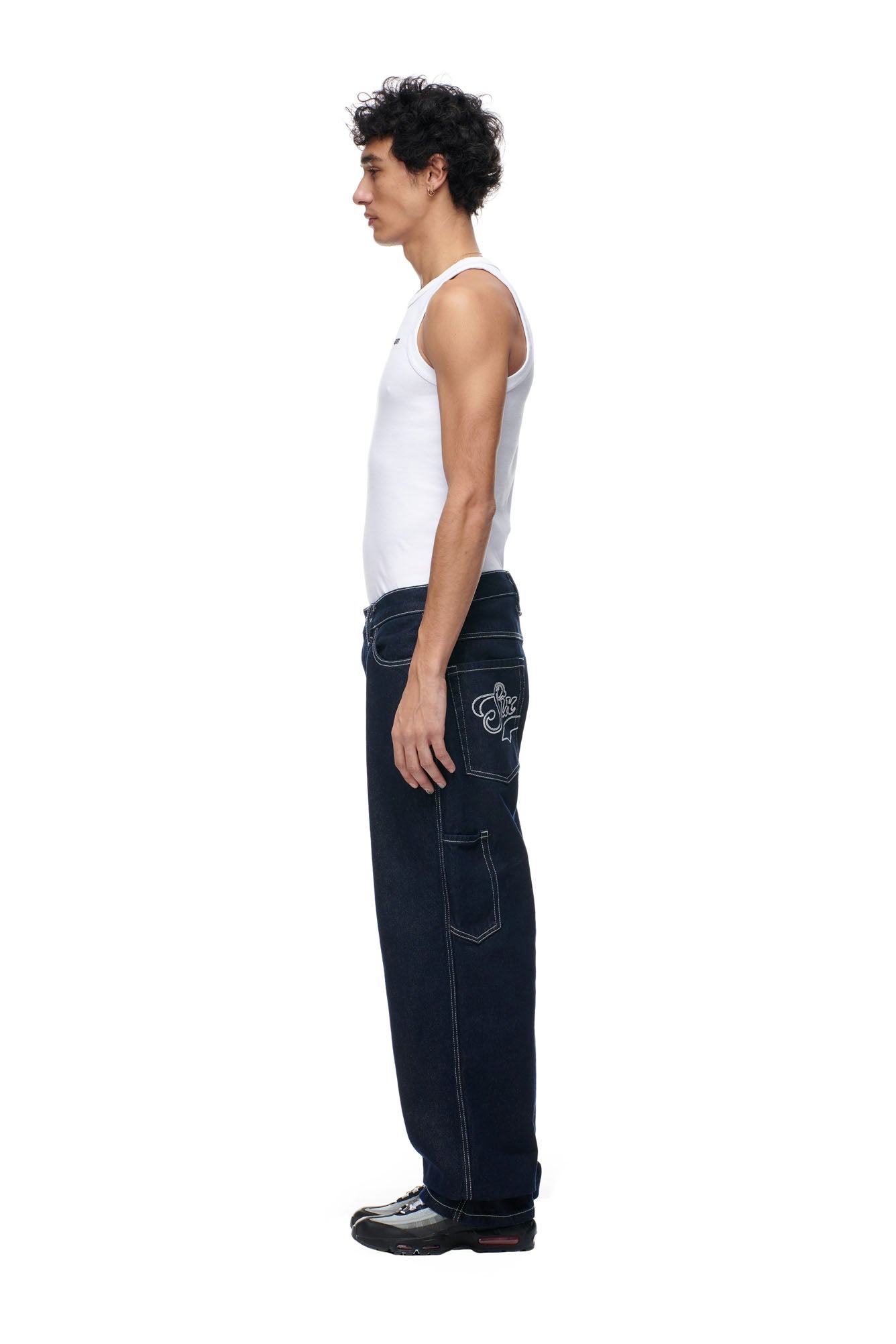 A man with curly hair stands in profile against a white background, wearing a white tank top, 6PM SCRIPT LOGO DENIM dark blue baggy jeans with large pockets and visible stitching, and black sneakers.