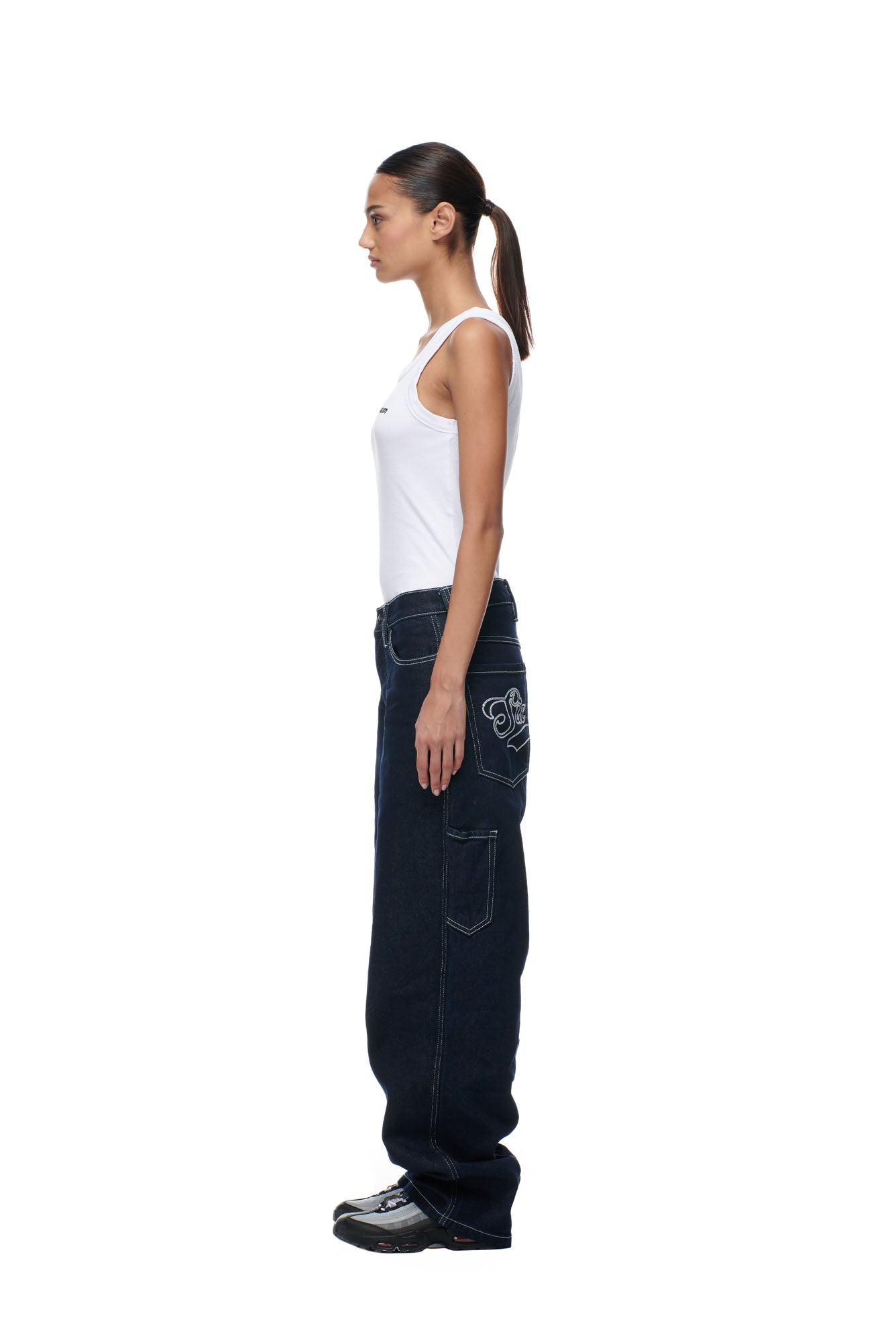 A woman with a ponytail stands in profile wearing a white tank top, black shoes, and 6PM SCRIPT LOGO DENIM—loose dark jeans with embroidered back pocket details—against a plain white background.