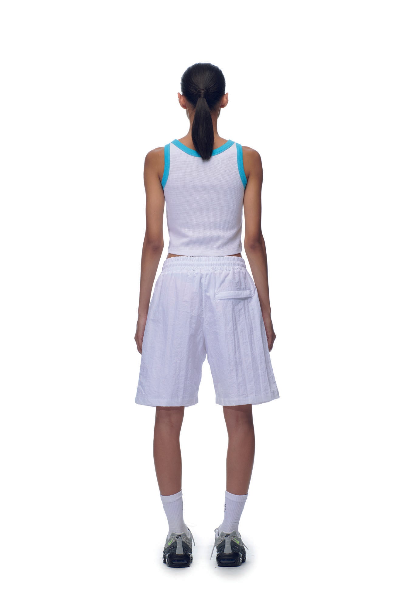 A person stands facing away against a plain white background, wearing the 6PM NH CROP TOP WHITE, loose white shorts, white socks, and black sneakers.