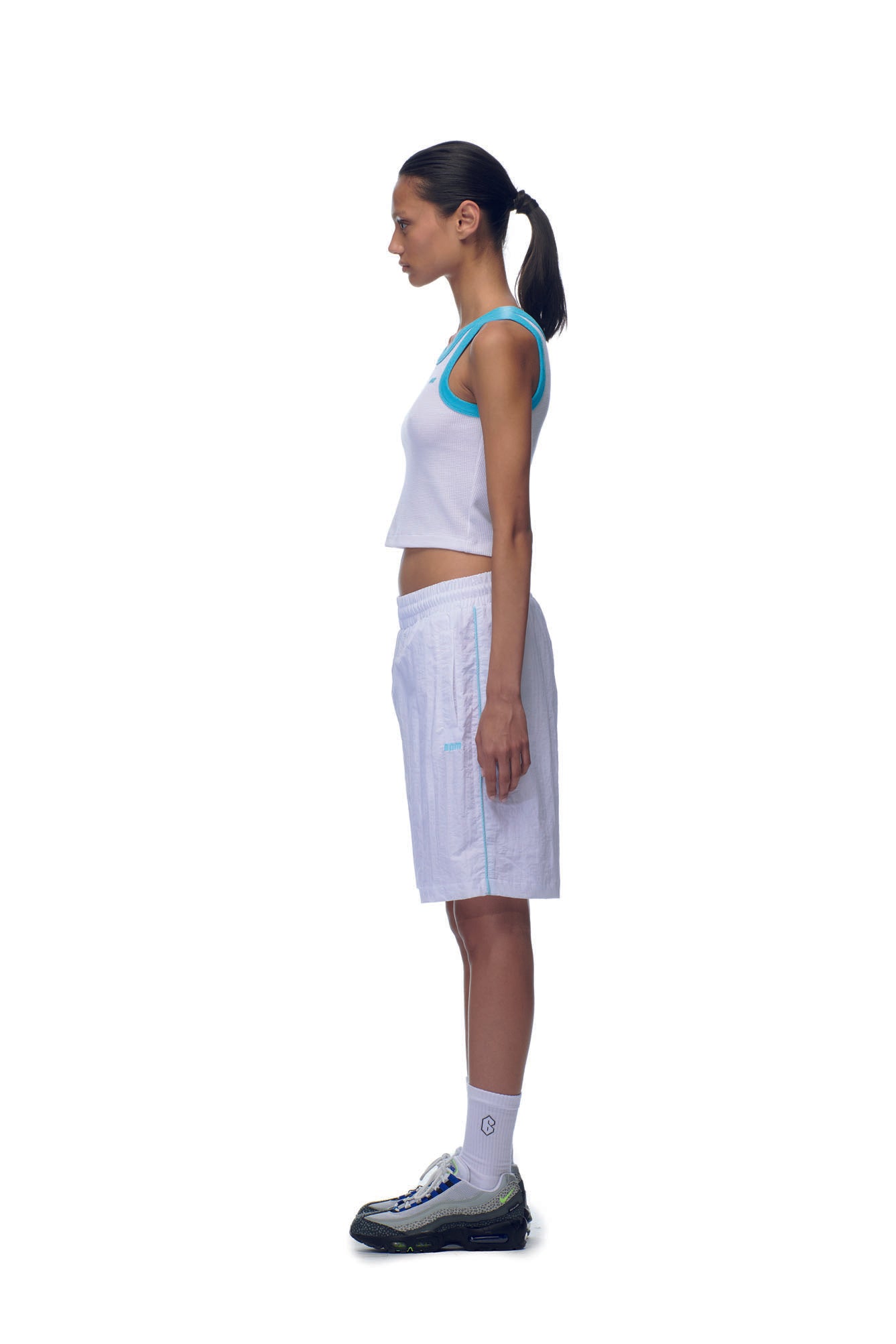 A person in profile stands against a white backdrop, wearing the 6PM NH CROP TOP WHITE, white shorts, white socks, and black-and-white sneakers, with their hair tied back in a ponytail.