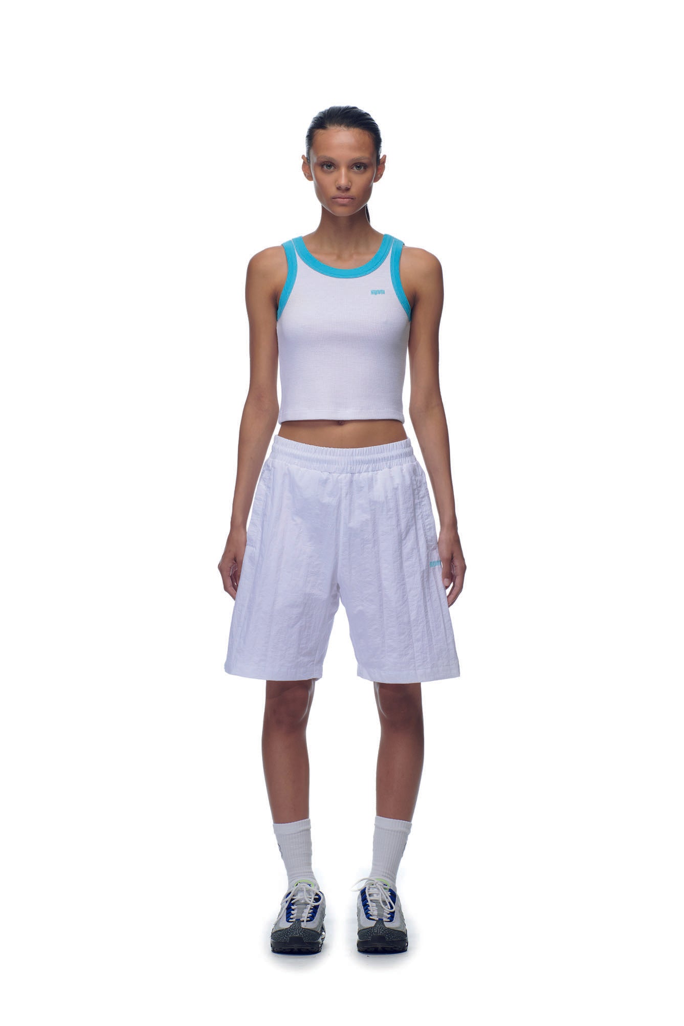 A person stands facing forward, wearing the 6PM NH CROP TOP WHITE with aqua blue trim, loose white knee-length shorts, white socks, and black-and-white sneakers against a plain white background.
