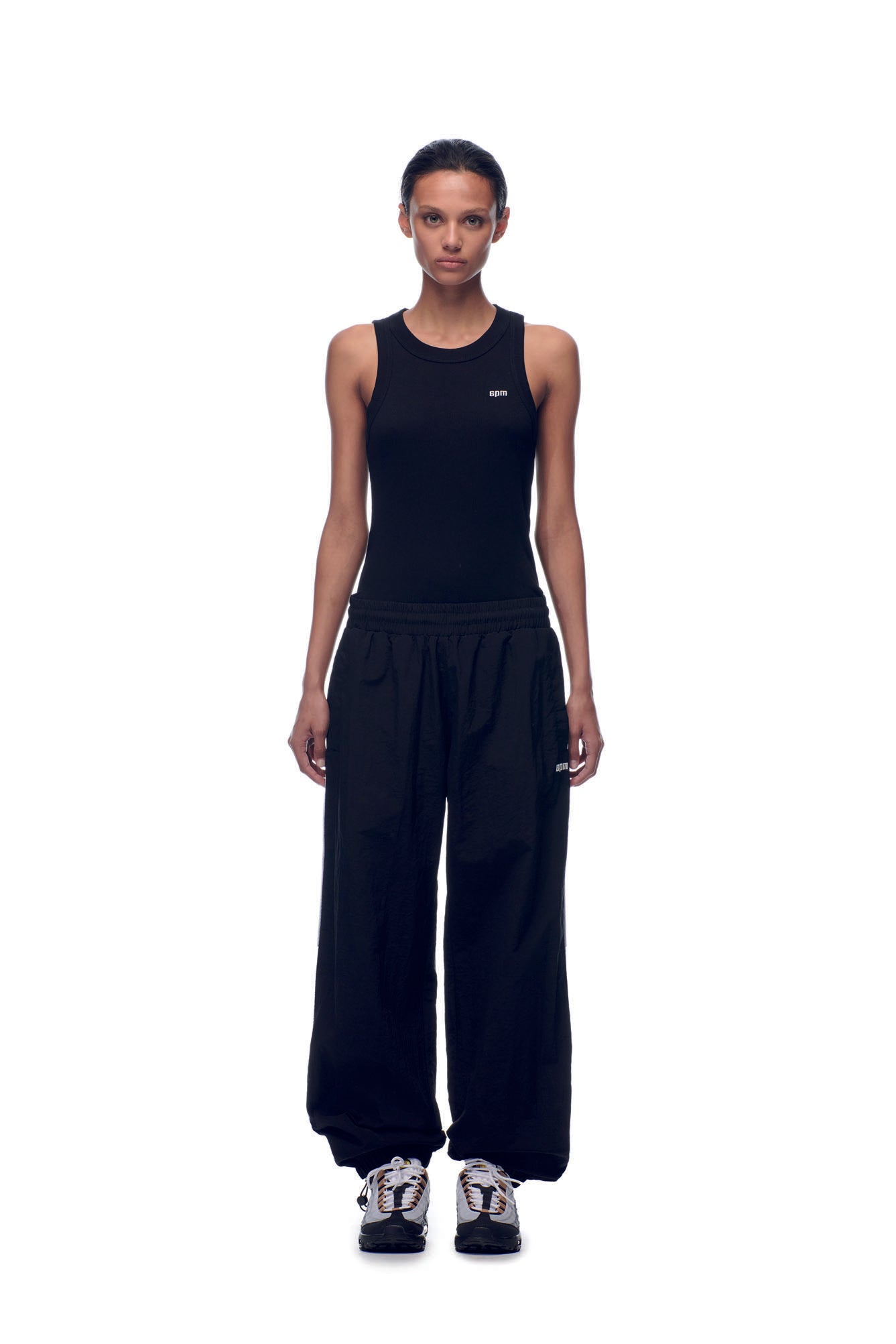 A person stands facing forward against a plain white background, wearing 6PM NH NYLON TRACKPANTS BLACK, a black sleeveless top, and white and black sneakers.