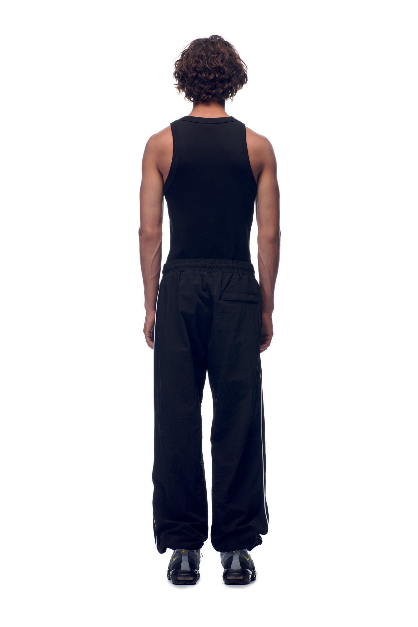 A person with curly hair faces away, wearing a black sleeveless top, black shoes, and 6PM NH NYLON TRACKPANTS BLACK against a white background.