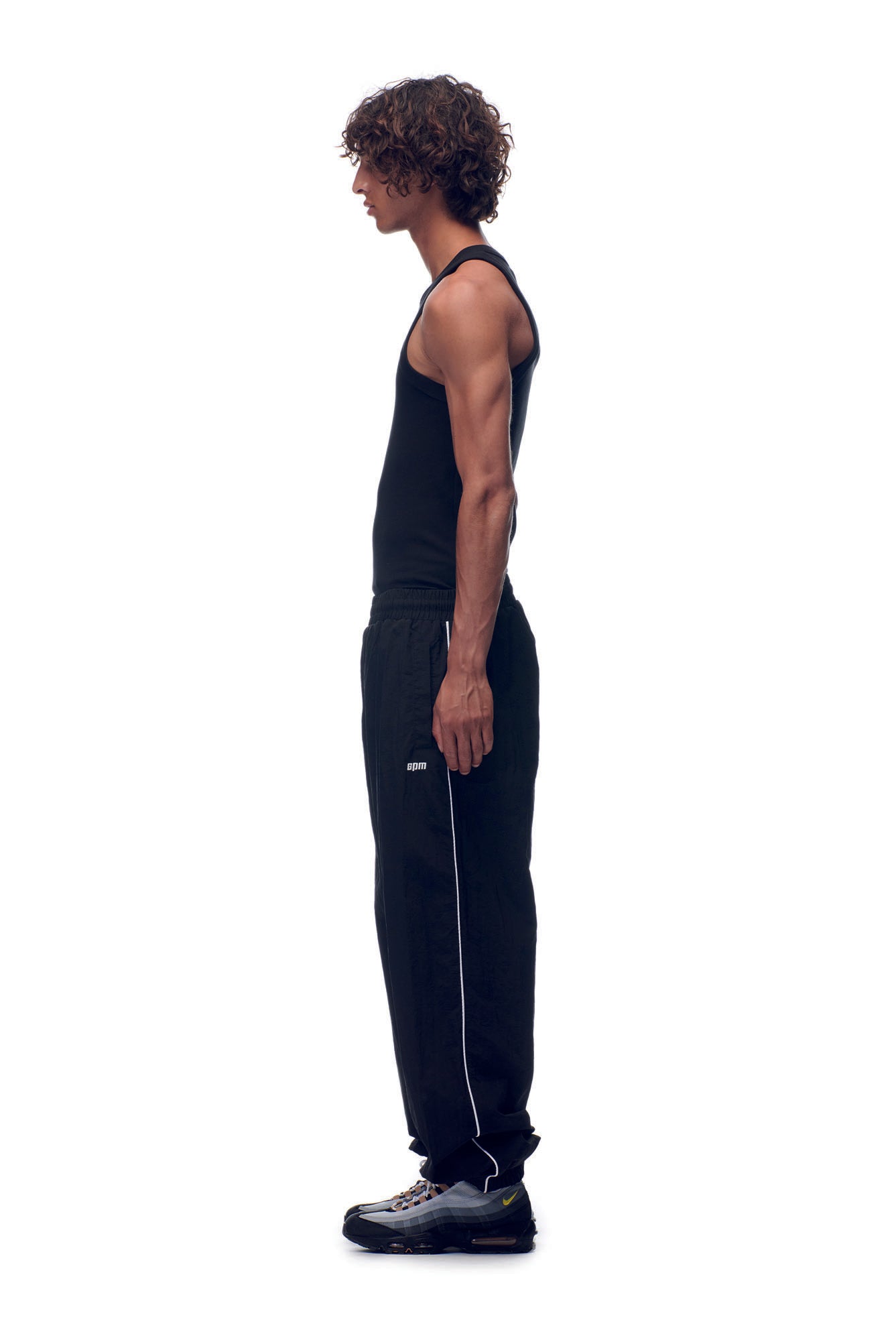 A young man with curly hair stands in profile against a white background, wearing 6PM NH NYLON TRACKPANTS BLACK with a black tank top and black sneakers.