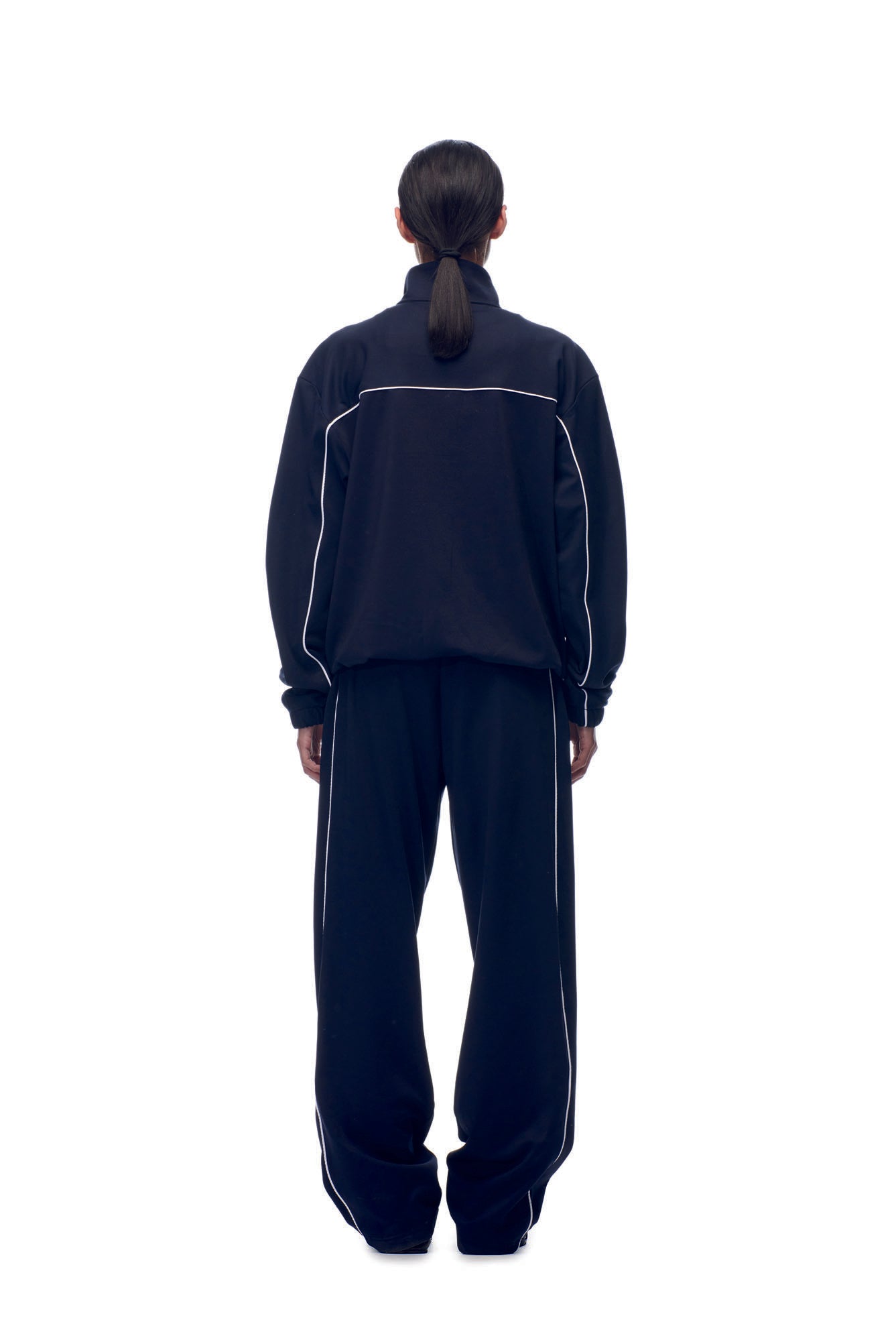 A person with long dark hair tied back stands facing away, wearing the 6PM TENNIS TRAINER JACKET BLUE, a loose-fitting tracksuit with white piping details, against a plain white background.