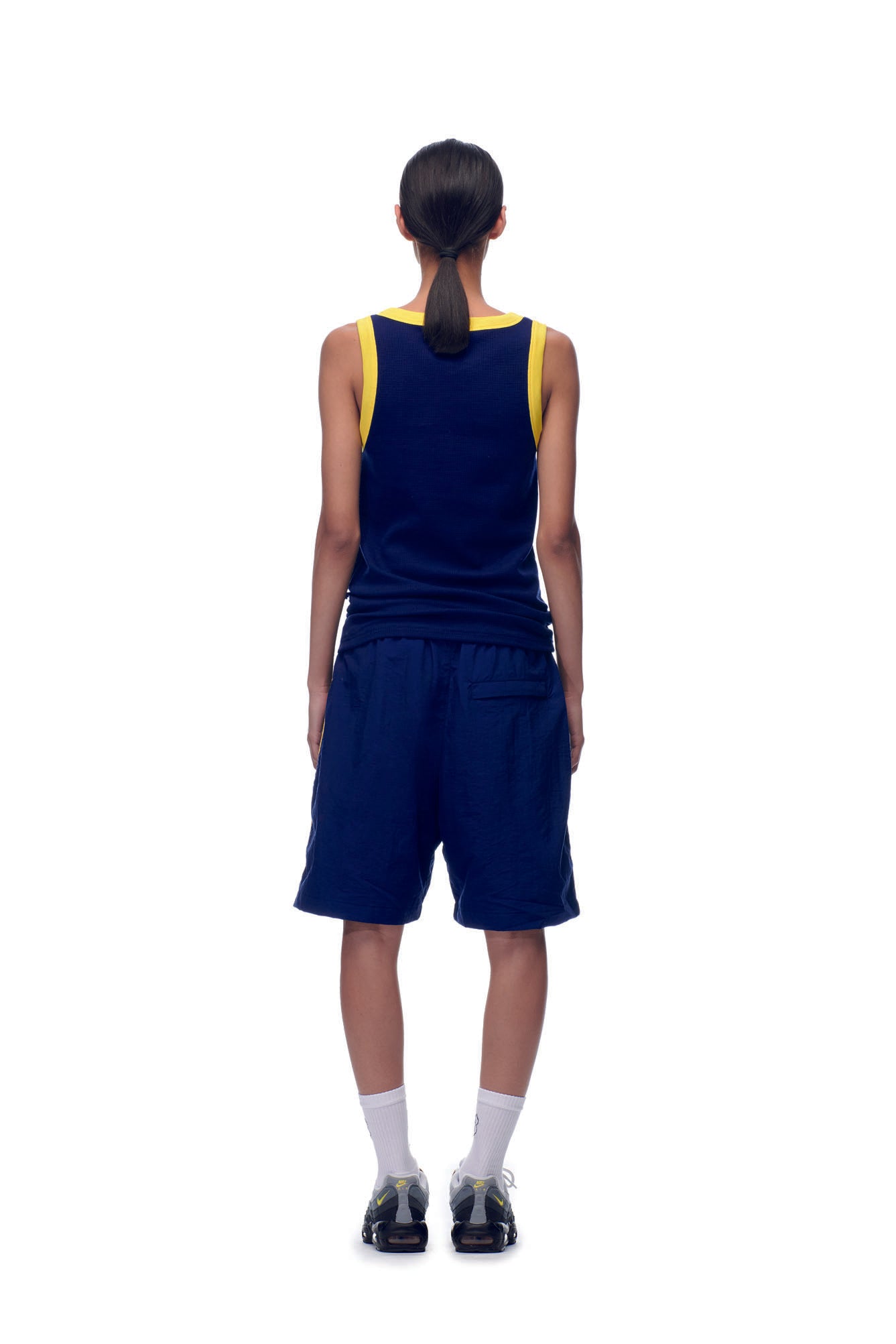 A person with their hair in a low ponytail stands against a white background, shown from behind, wearing the 6PM NH TANKTOP NAVY/YELLOW, navy shorts, white socks, and black athletic shoes.