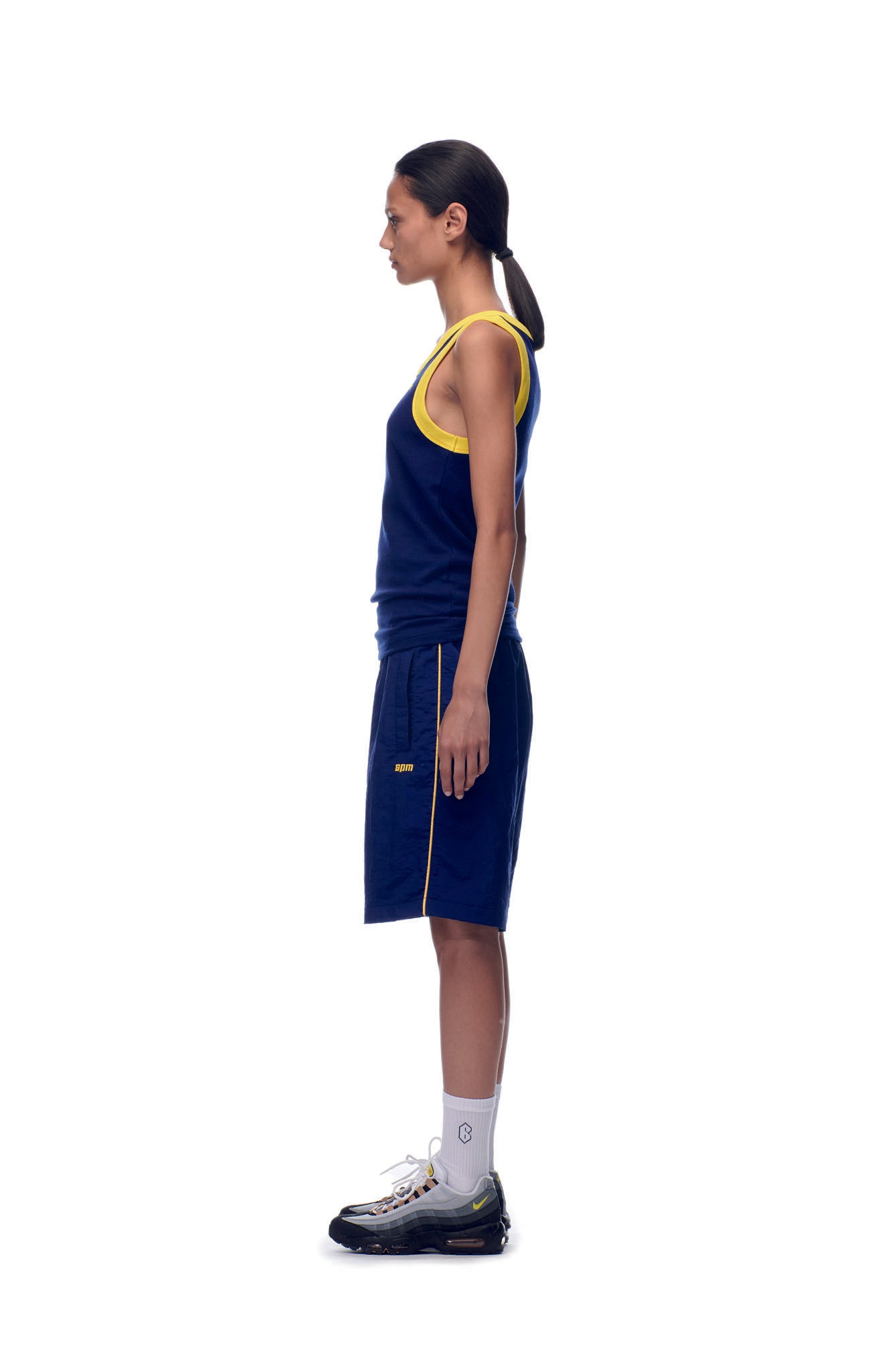 A person stands in profile against a white background, wearing the 6PM NH TANKTOP NAVY/YELLOW, matching shorts with yellow accents, white socks, black athletic shoes, and their hair in a low ponytail.