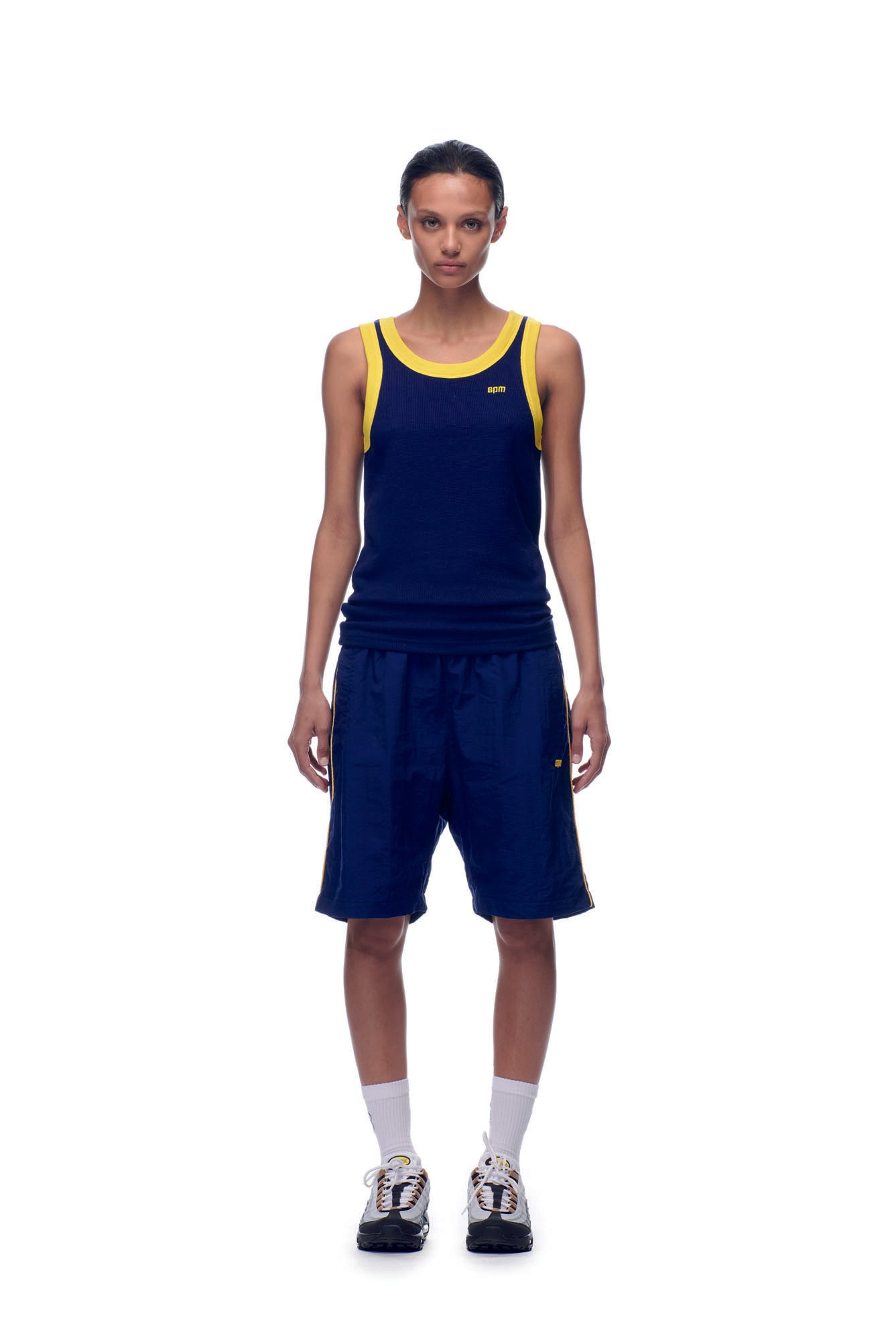 A person stands facing forward against a plain white background, wearing the 6PM NH TANKTOP NAVY/YELLOW, navy athletic shorts, white socks, and black and white sneakers.