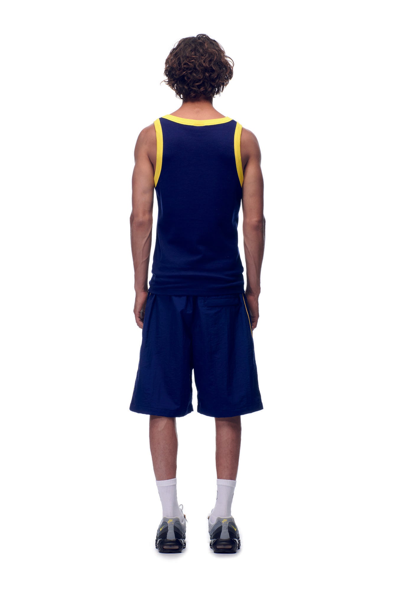 A person with curly hair faces away, wearing the 6PM NH TANKTOP NAVY/YELLOW, navy shorts, white socks, and black sneakers, set against a white background.