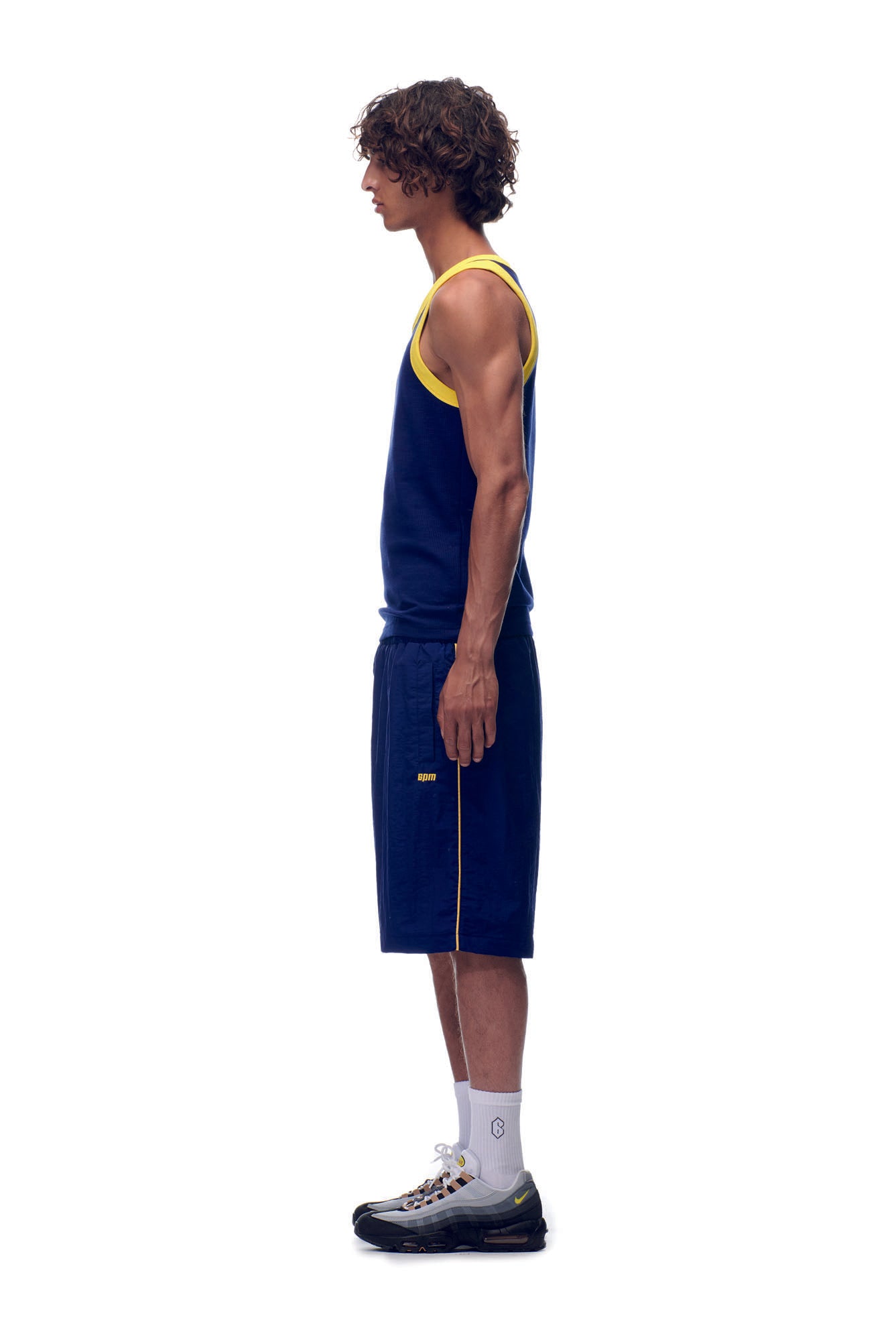 A man in profile stands against a white background, wearing the 6PM NH TANKTOP NAVY/YELLOW, matching navy shorts, white socks, and black sneakers.
