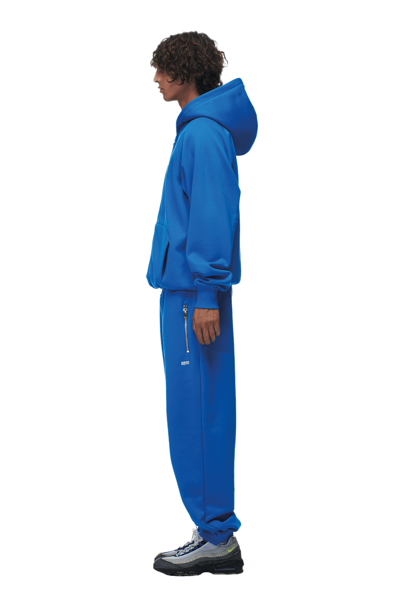 A person in profile wears the 6PM ZIP HOODIE PRINCESS BLUE with matching blue sweatpants featuring zipper pockets, styled for everyday wear. Chunky black and gray sneakers finish the outfit. Background is transparent.