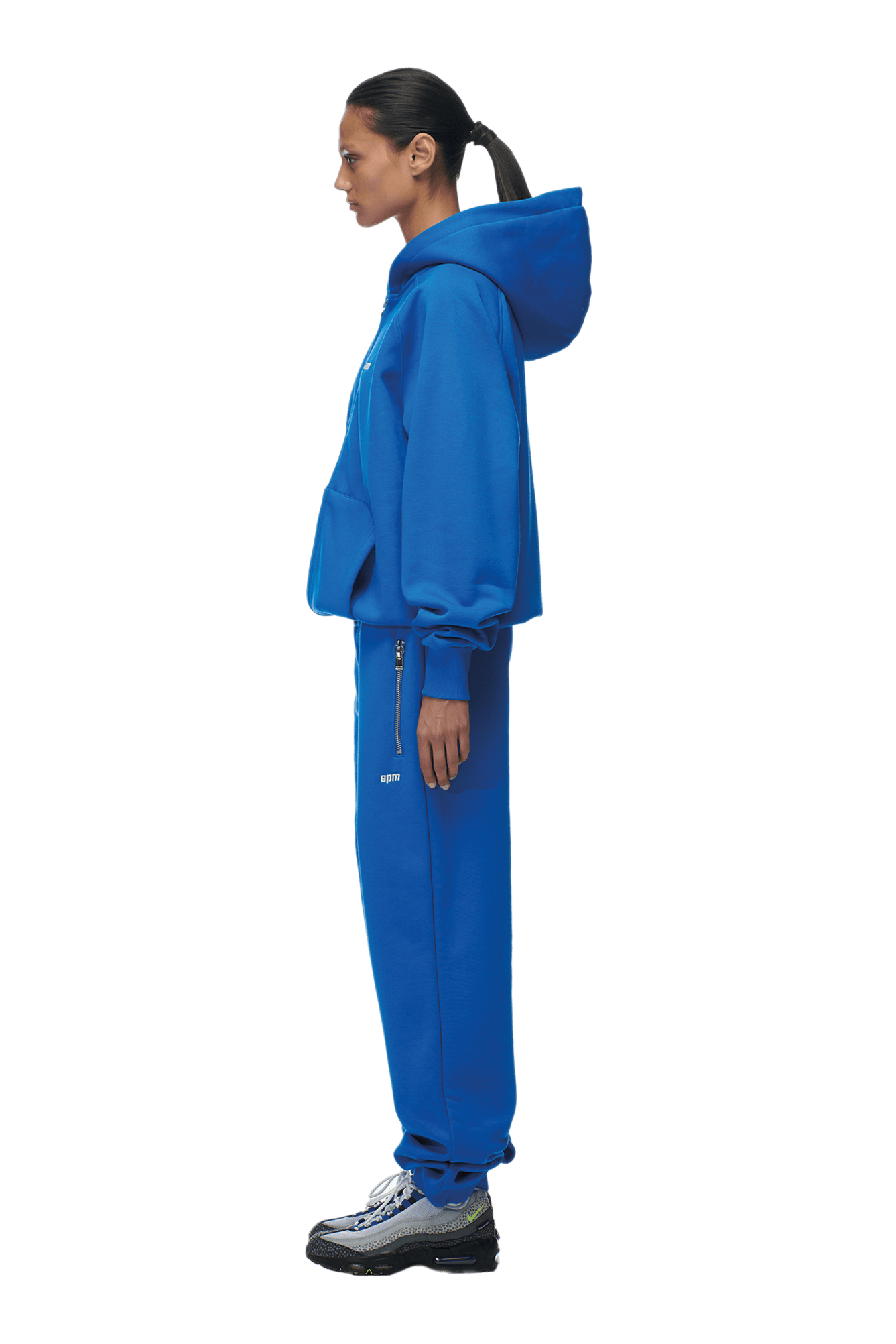 A woman in profile wears the 6PM ZIP HOODIE PRINCESS BLUE with matching sweatpants and black-and-white sneakers. Her hair is tied back in a low ponytail as she looks straight ahead against a white background.
