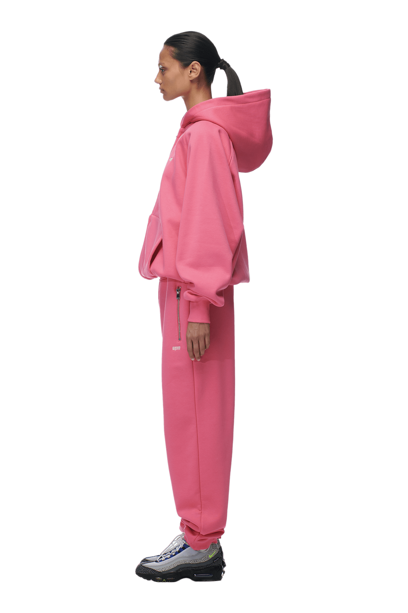 A woman in profile wears the 6PM ZIP HOODIE HOT PINK with matching sweatpants and gray sneakers; her dark hair is tied back. The outfit’s clean finish is perfect for everyday wear. Background is transparent.