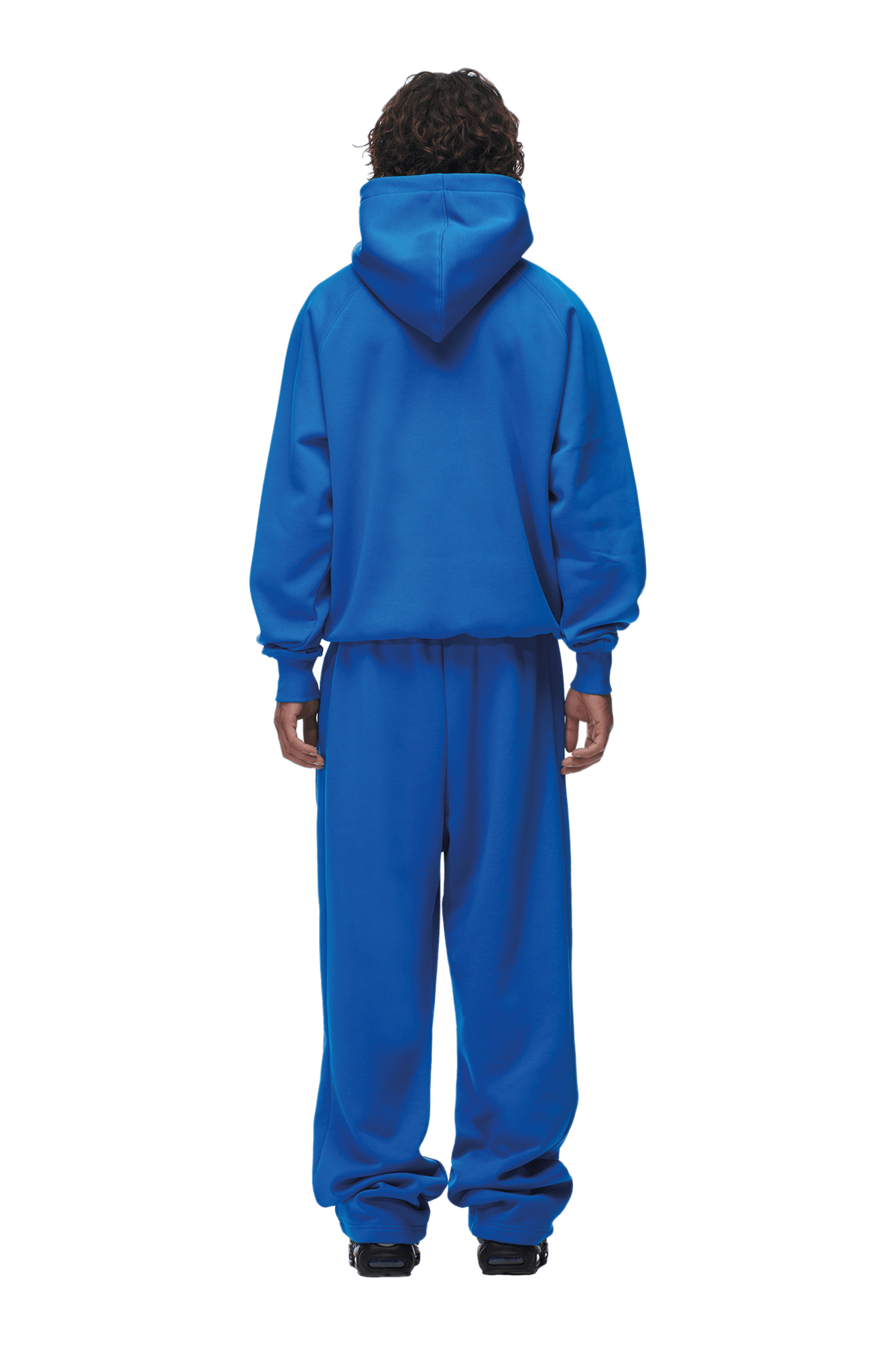 A person with curly hair faces away, wearing the 6PM HOODIE PRINCESS BLUE with raglan sleeves and hood up, paired with matching sweatpants. The loose fit and classic kangaroo pocket make this set ideal for everyday style against a plain background.