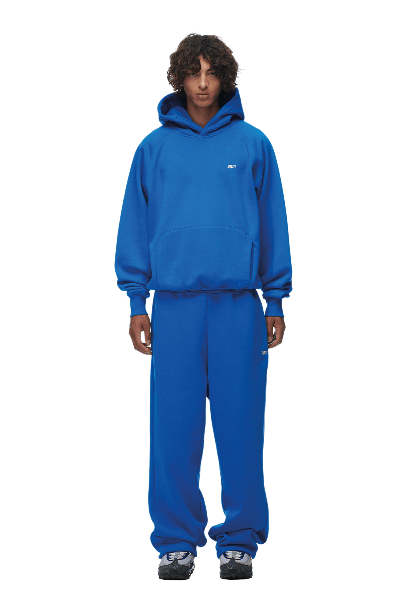 A person with curly hair models the 6PM HOODIE PRINCESS BLUE and matching sweatpants, featuring raglan sleeves and a kangaroo pocket, paired with black and white sneakers—ideal for everyday wear against a transparent background.