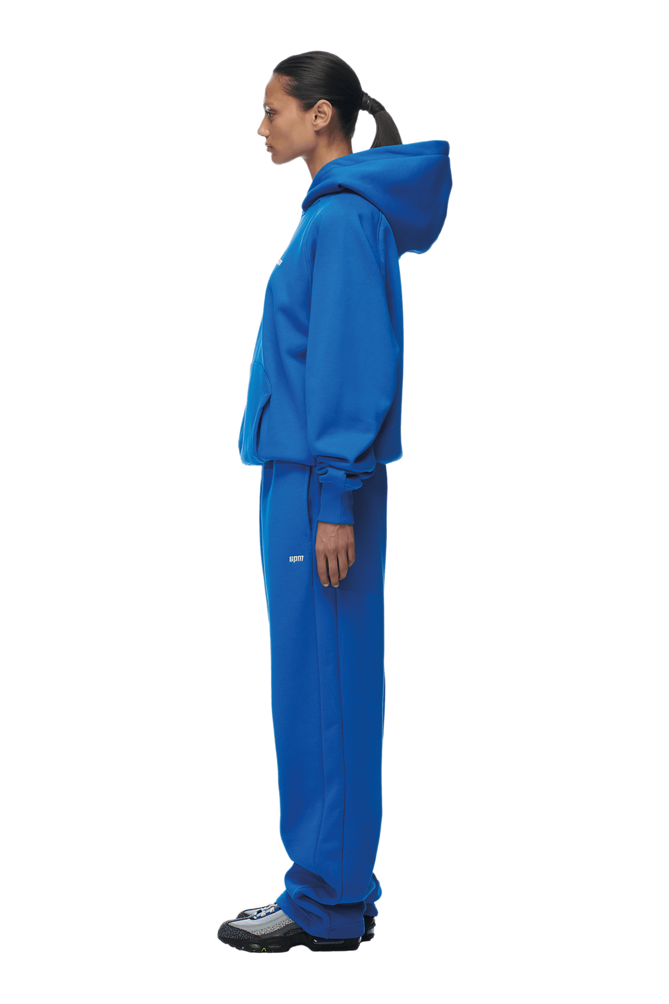 A woman stands in profile against a transparent background, wearing the 6PM HOODIE PRINCESS BLUE raglan-sleeved hoodie and sweatpants set with a kangaroo pocket, black sneakers, and her hair in a ponytail—ideal for everyday wear.