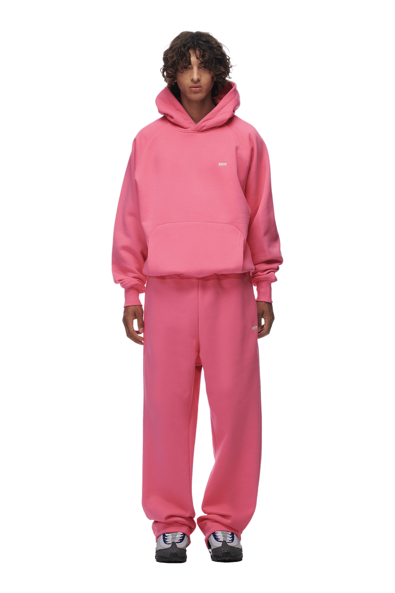 A person with curly hair models the 6PM HOODIE HOT PINK, a relaxed raglan-sleeve hoodie with a kangaroo pocket and matching sweatpants, styled with black and white sneakers—ideal for everyday wear against a plain backdrop.