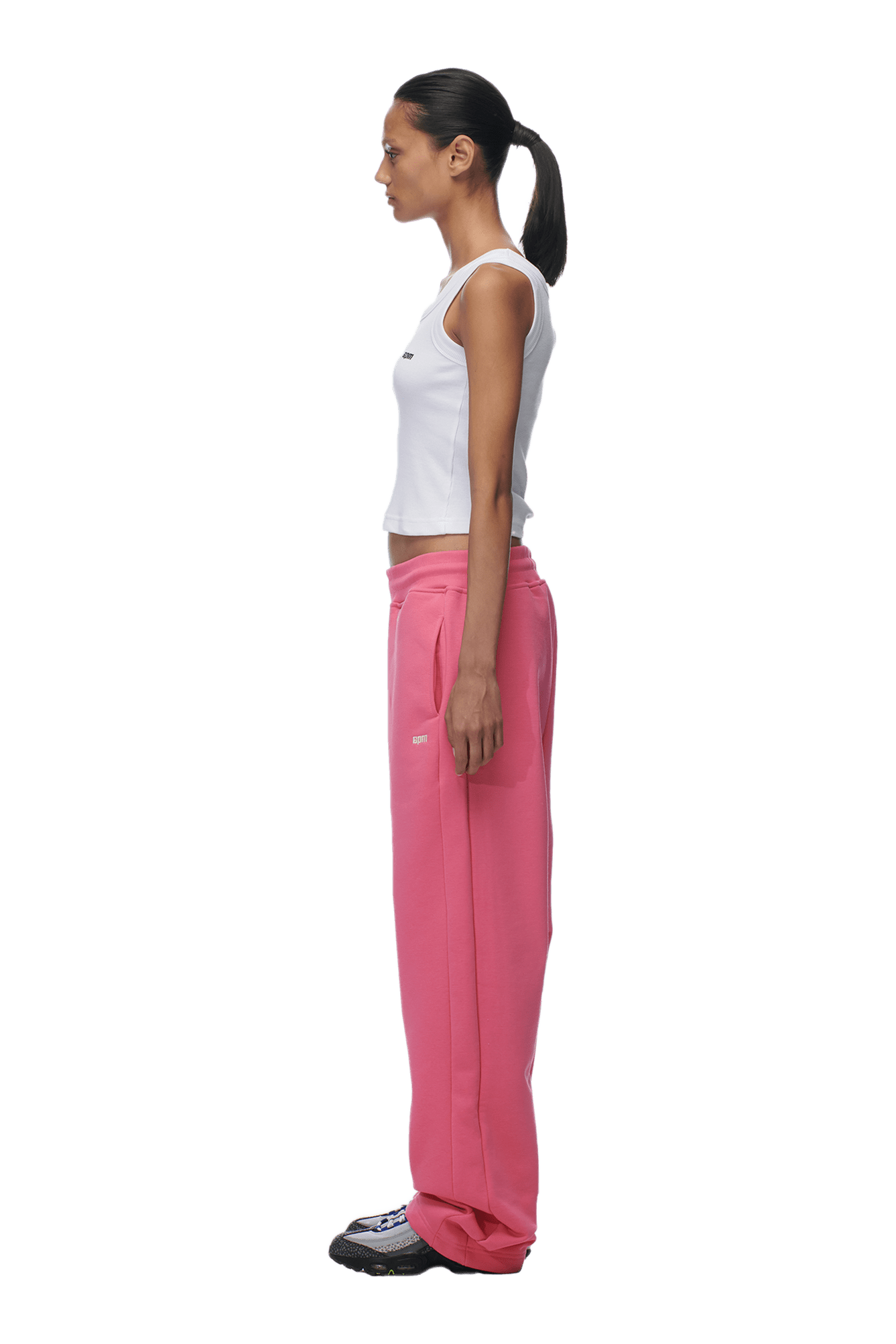 A woman stands in profile, wearing a white sleeveless top and 6PM’s OPEN LEG JOGGER HOT PINK sweatpants with black sneakers. Her dark hair is tied in a ponytail as she faces left against a plain background.